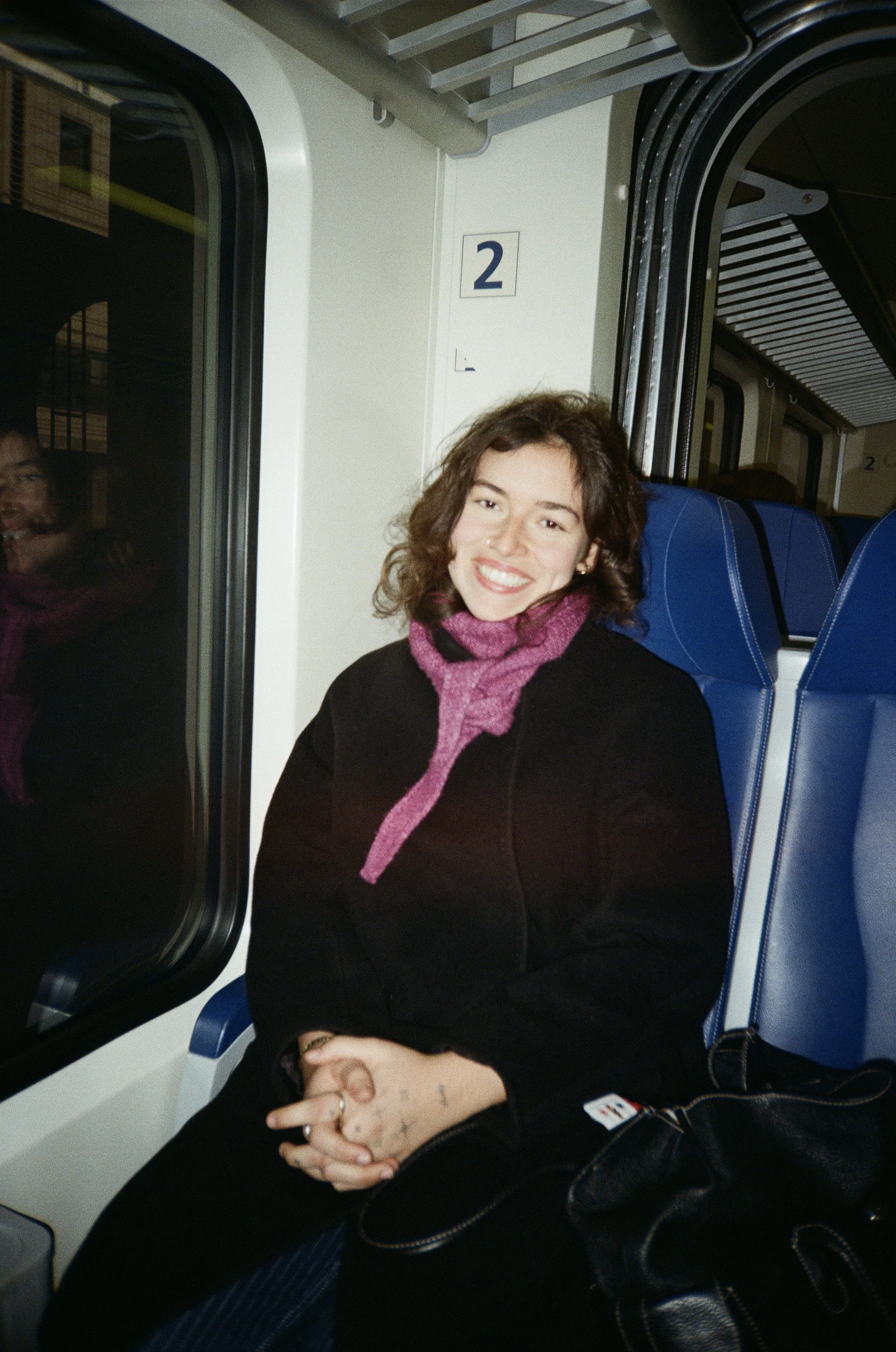 A woman with curly brown hair and a pink scarf smiling while sitting on a train, with a window to her left and a blue seat behind her.