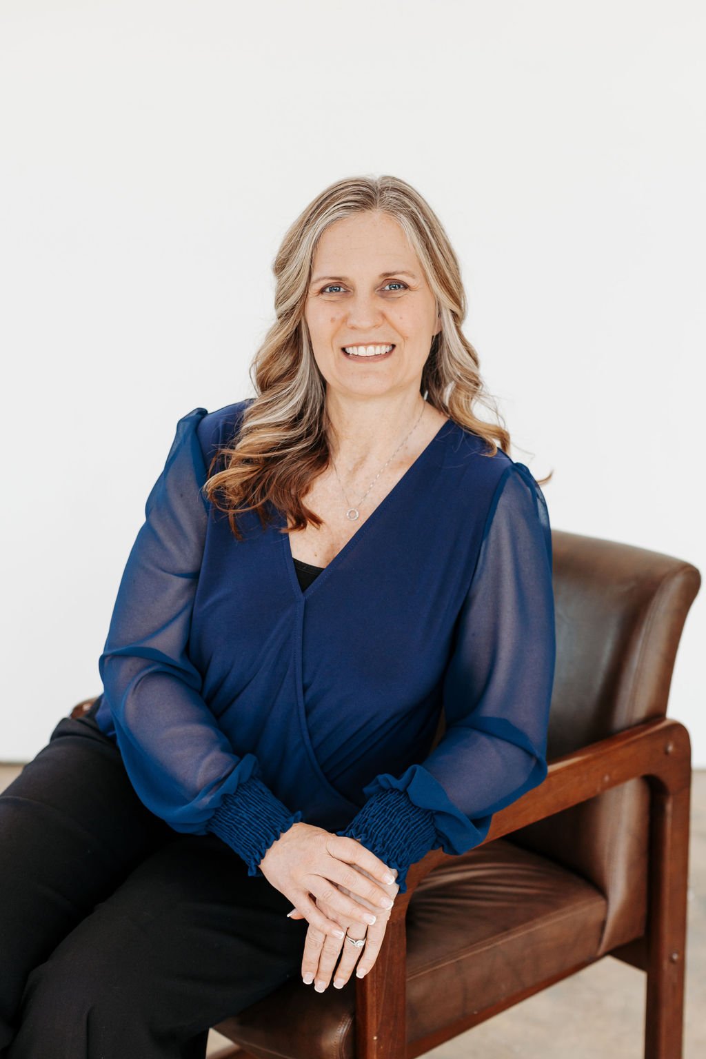 A woman with wavy, shoulder-length blonde hair, wearing a navy blue blouse and black pants, sitting on a wooden chair against a plain white background, smiling at the camera.
