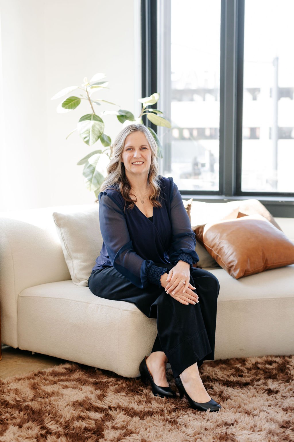 A woman with wavy gray hair sitting on a cream-colored sofa by a large window, with a green plant and a brown pillow behind her, indoors with natural light.