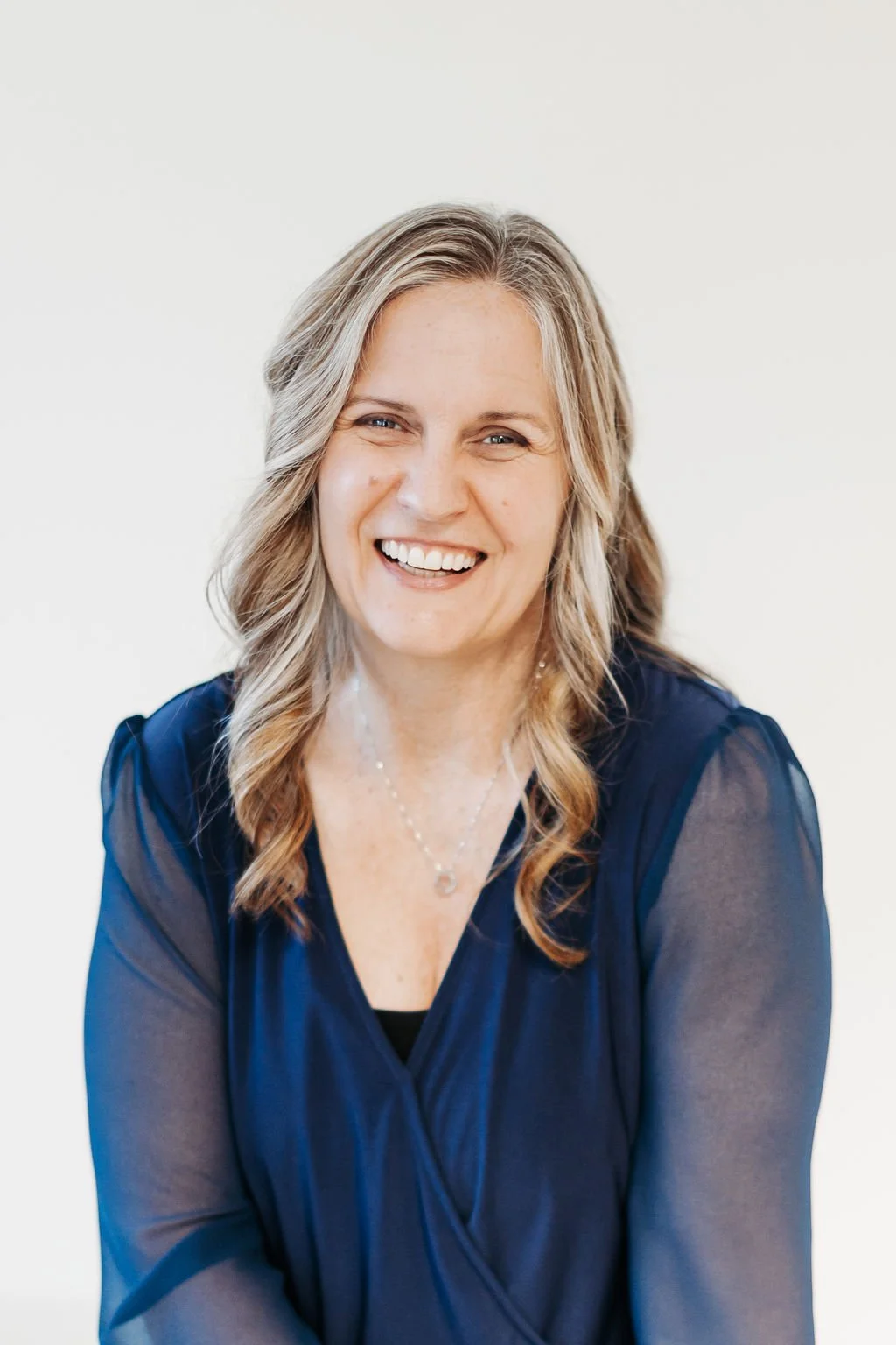 A woman with wavy blonde hair smiling, wearing a navy blue sheer blouse and a silver necklace, against a plain white background.