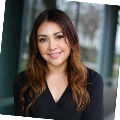 A woman with long brown hair and a black top smiling indoors near a window.