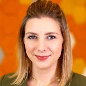 A young woman with blonde hair, wearing a green top, smiling at the camera with an orange background.