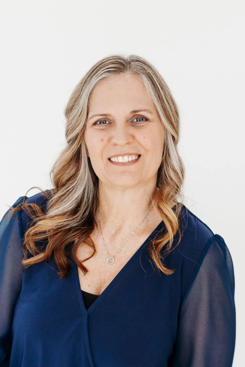 A woman with long, wavy blonde hair and blue eyes, wearing a navy blue top and a silver necklace, smiling against a plain white background.
