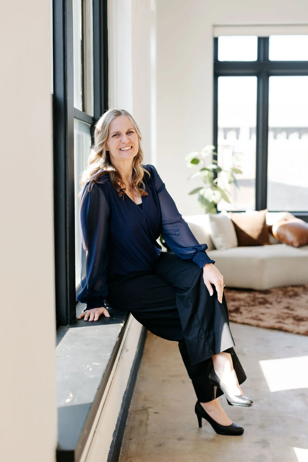 A woman sitting on a windowsill, wearing a navy blouse, black pants, and black high heels, smiling indoors with a beige sofa, pillows, and a large window in the background.