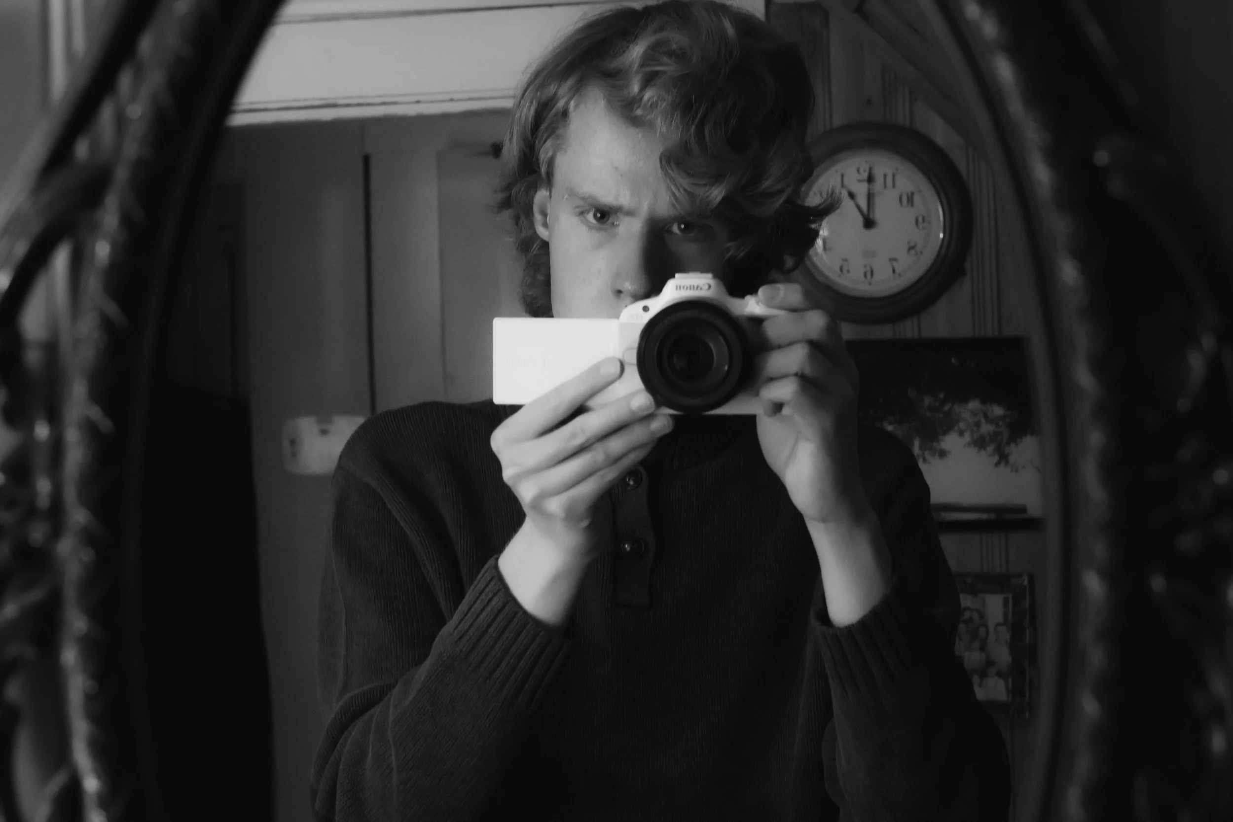 A person taking a mirror selfie with a white camera, with a clock on the wall in the background showing 11:25.