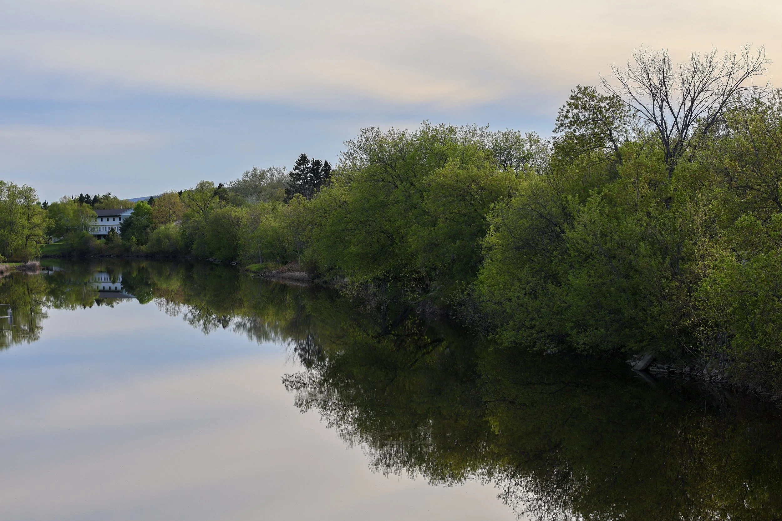 A peaceful river reflecting surrounding trees and a house with a white exterior and dark roof, under a cloudy sky.