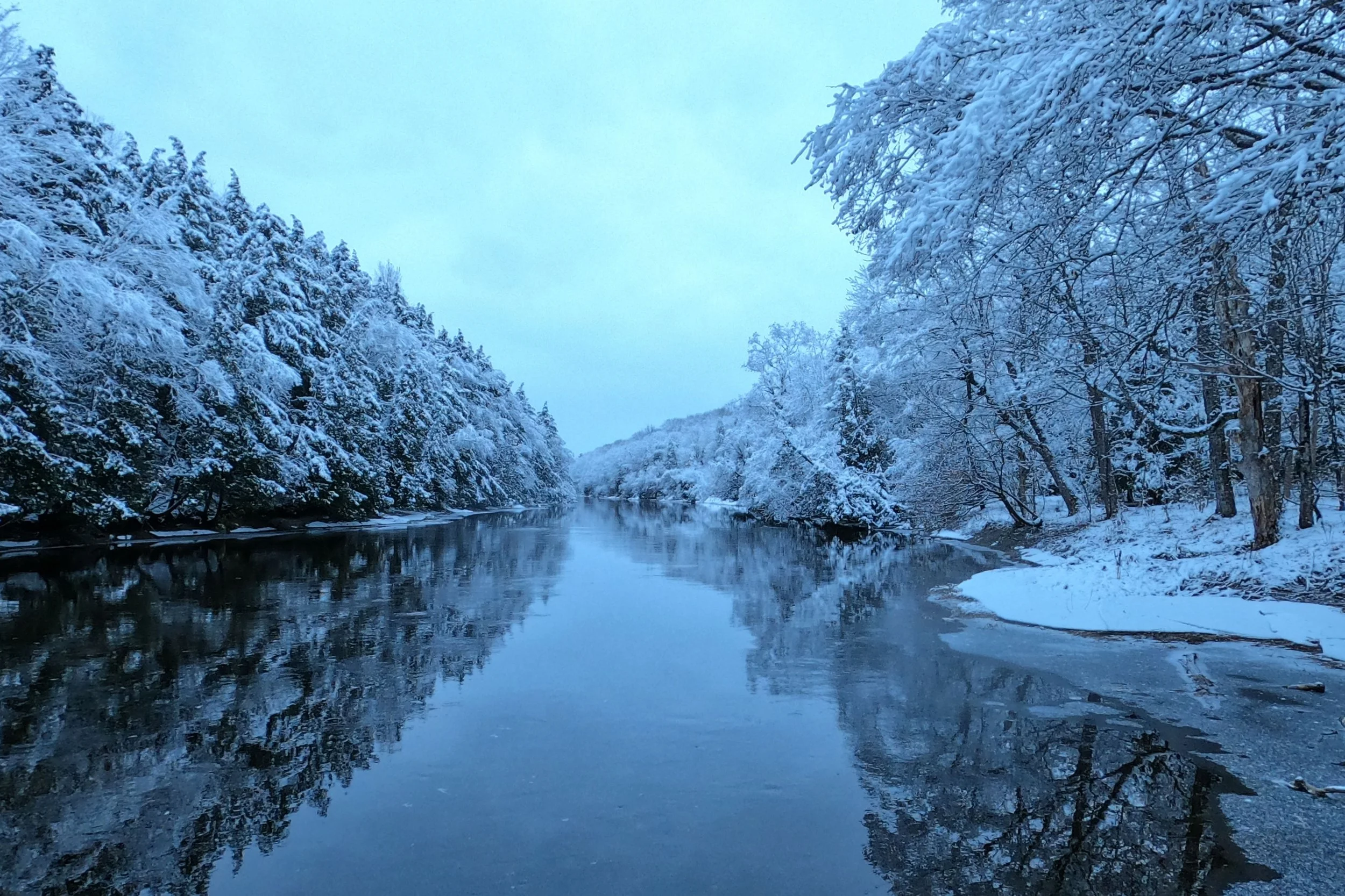 A river surrounded by snow-covered trees on both sides, with the water reflecting the snowy landscape and a cloudy sky.