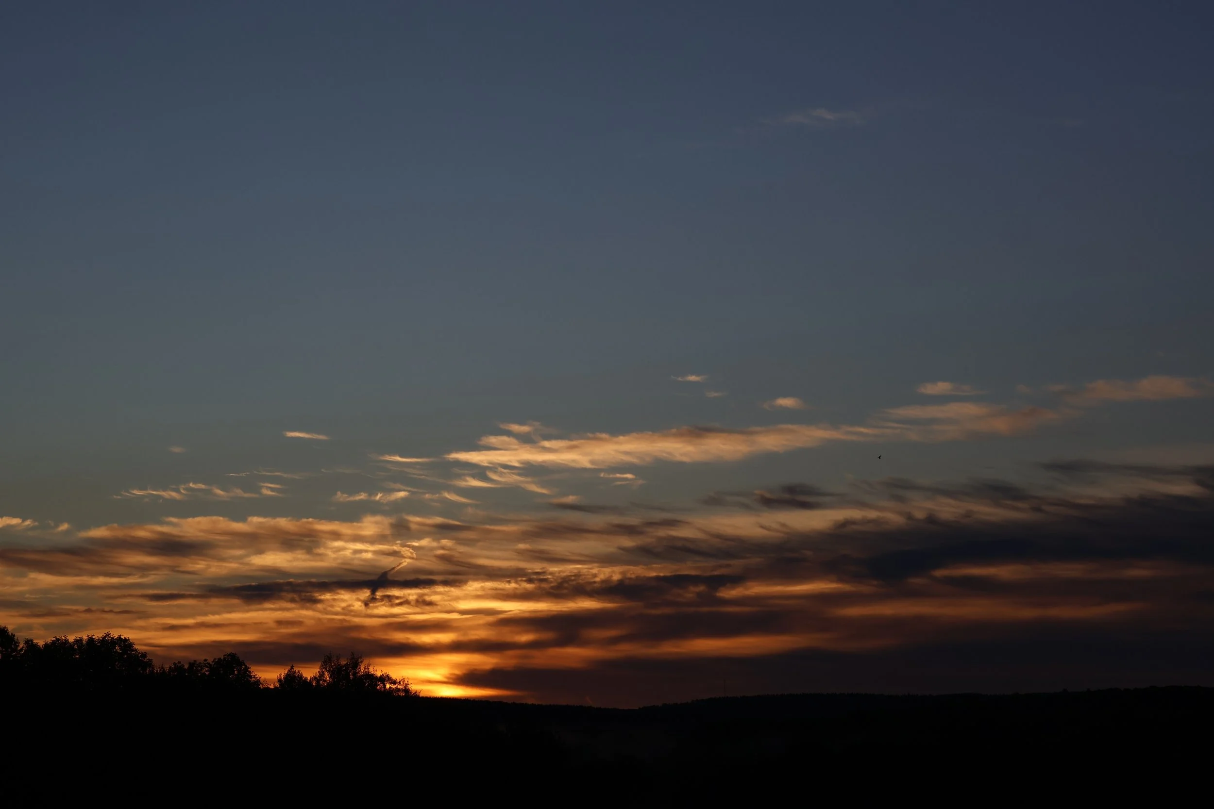 Sunset with orange and yellow clouds casting a glow over a silhouette of trees and landscape.