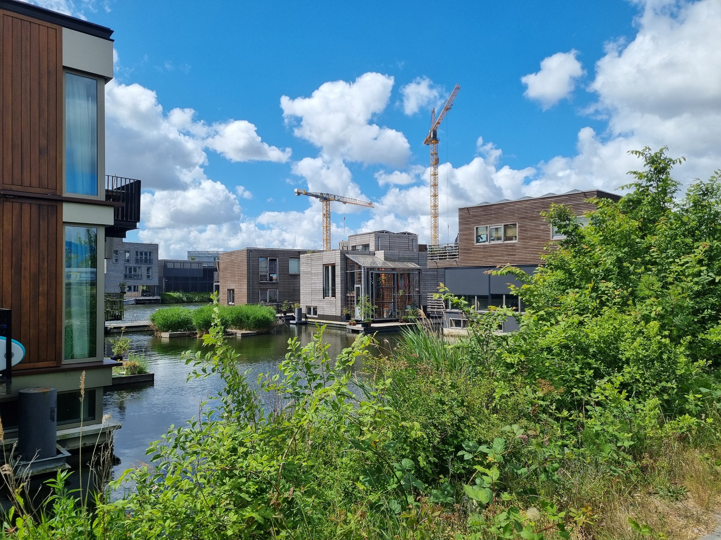 Modern residential buildings with wooden and concrete facades beside a waterway, under a blue sky with scattered clouds, and construction cranes in the background.