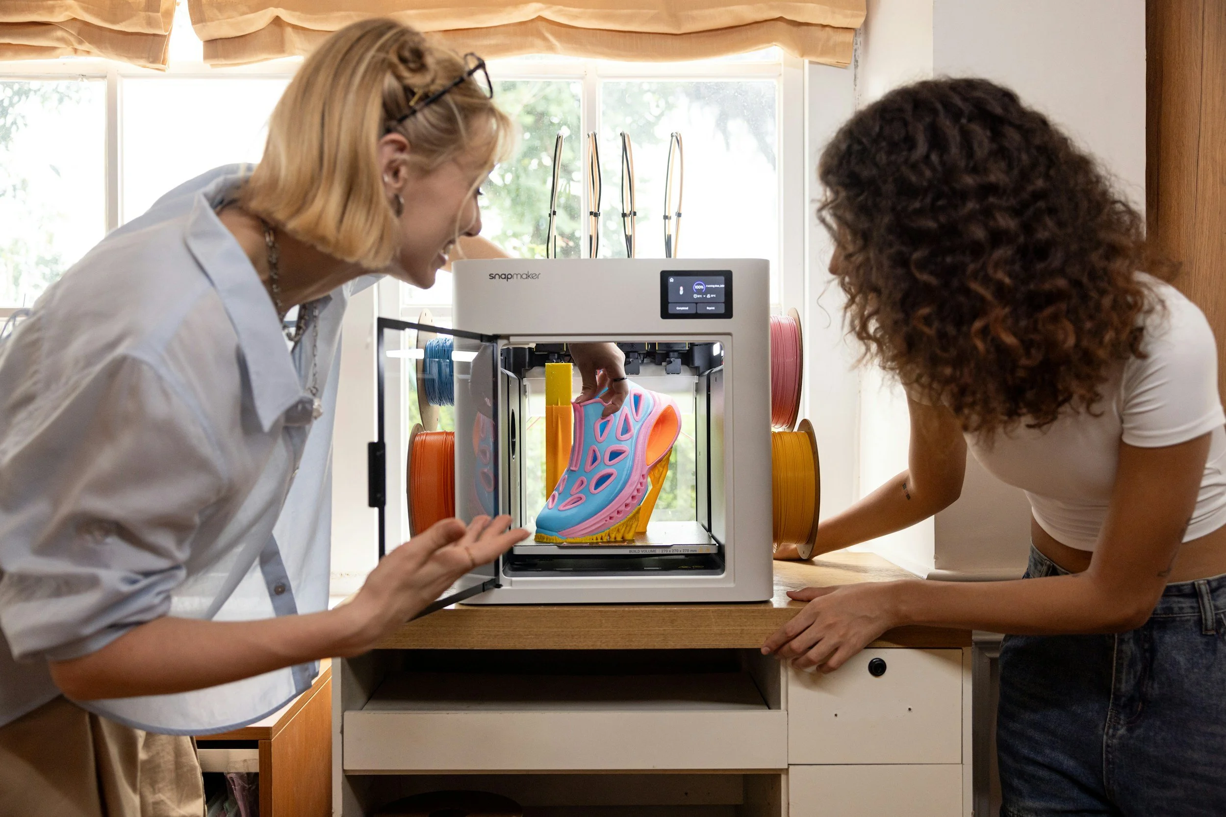 Two women are examining a 3D printed shoe model inside a 3D printer with spool filaments mounted inside. The women are standing in a room with large windows and wooden furniture.