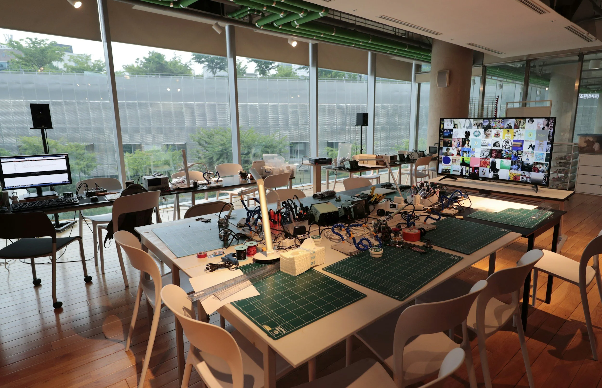 A large indoor workspace with tables covered in electronics, tools, and equipment, next to floor-to-ceiling windows showing trees outside.
