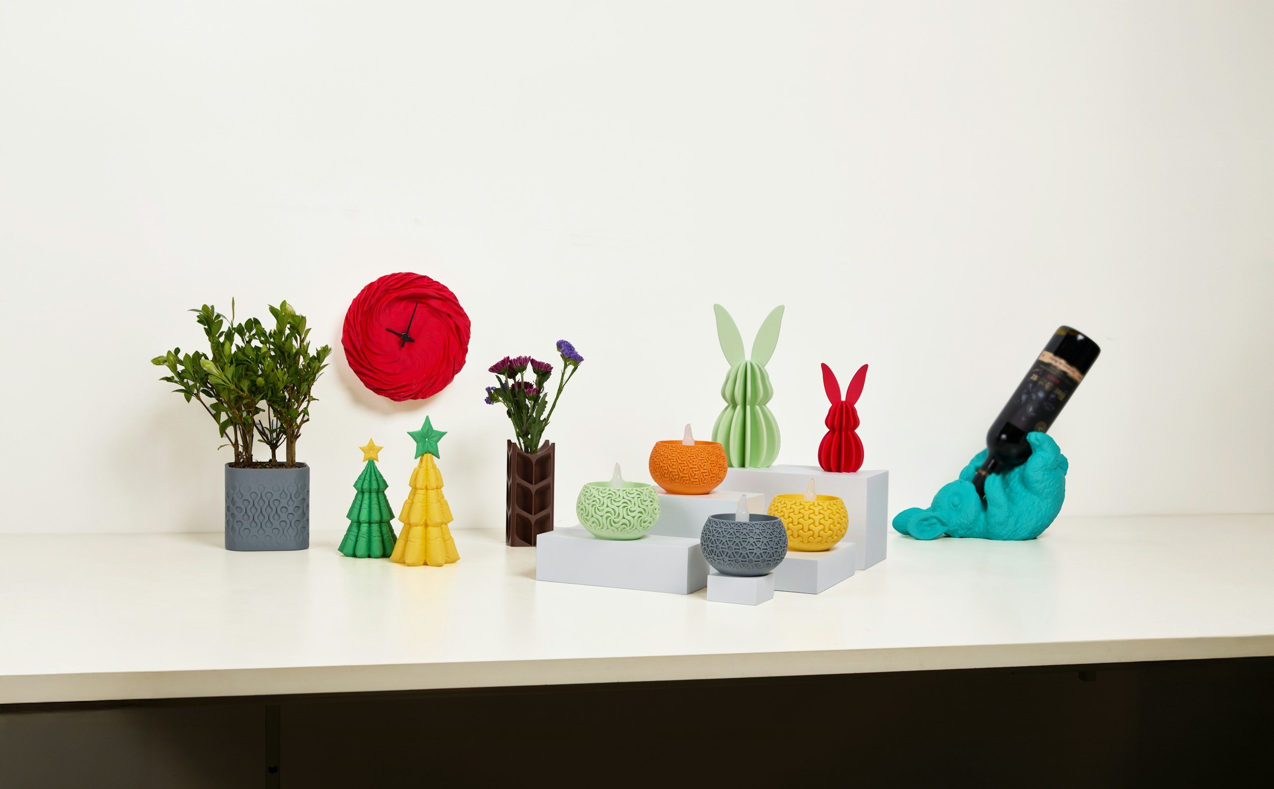 Arrangement of decorative items including potted green plant, red paper flower, small colorful Christmas trees, candles in patterned holders, rabbit-shaped ornaments, and a person wearing blue gloves holding a handheld device on a white table against a plain background.