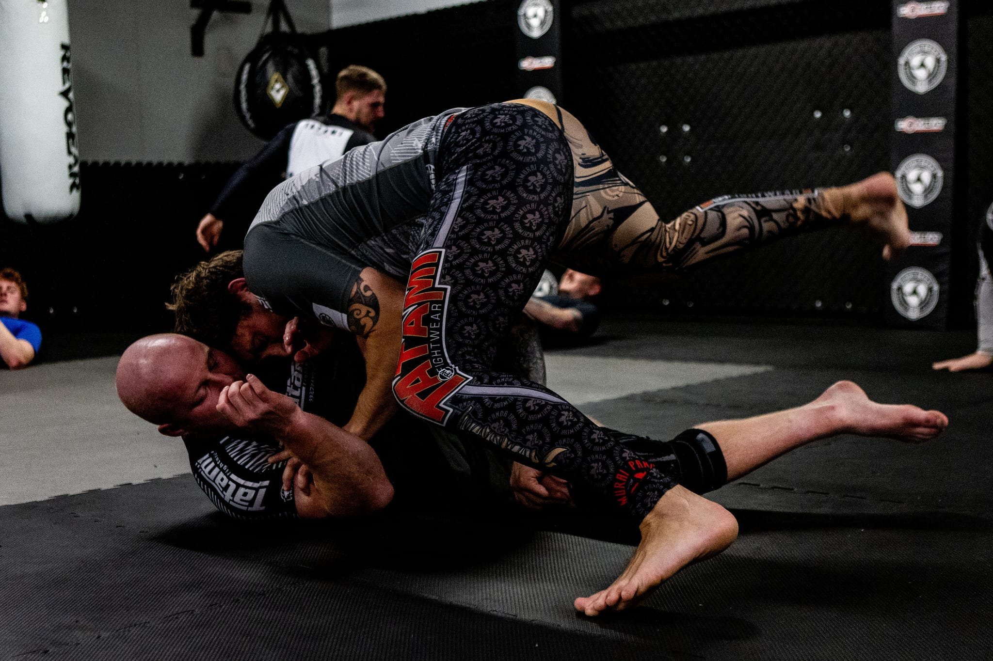 Two men practicing Brazilian Jiu-Jitsu on a black mat in a gym. One man is on top, while the other is on the bottom in a grappling position. Several children and a punching bag are visible in the background.