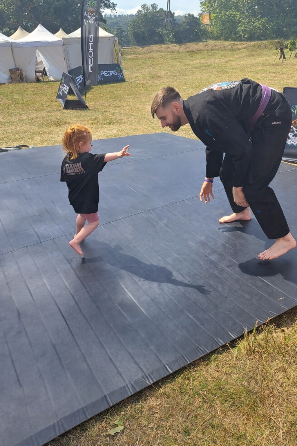 A young girl and an adult man are practicing Brazilian Jiu-Jitsu outdoors on a black mats. The girl is standing on one foot with outstretched arms, while the man is bent over in a martial arts stance. They are surrounded by a grass field with tents and banners in the background.
