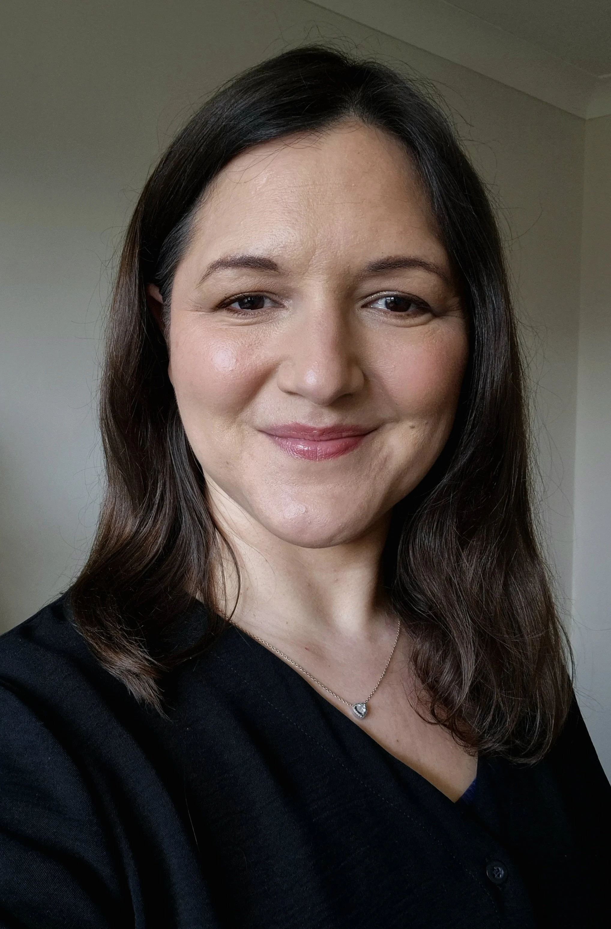 A woman with dark brown hair, wearing a black top and a silver necklace with a heart-shaped pendant, smiling in front of a plain wall.
