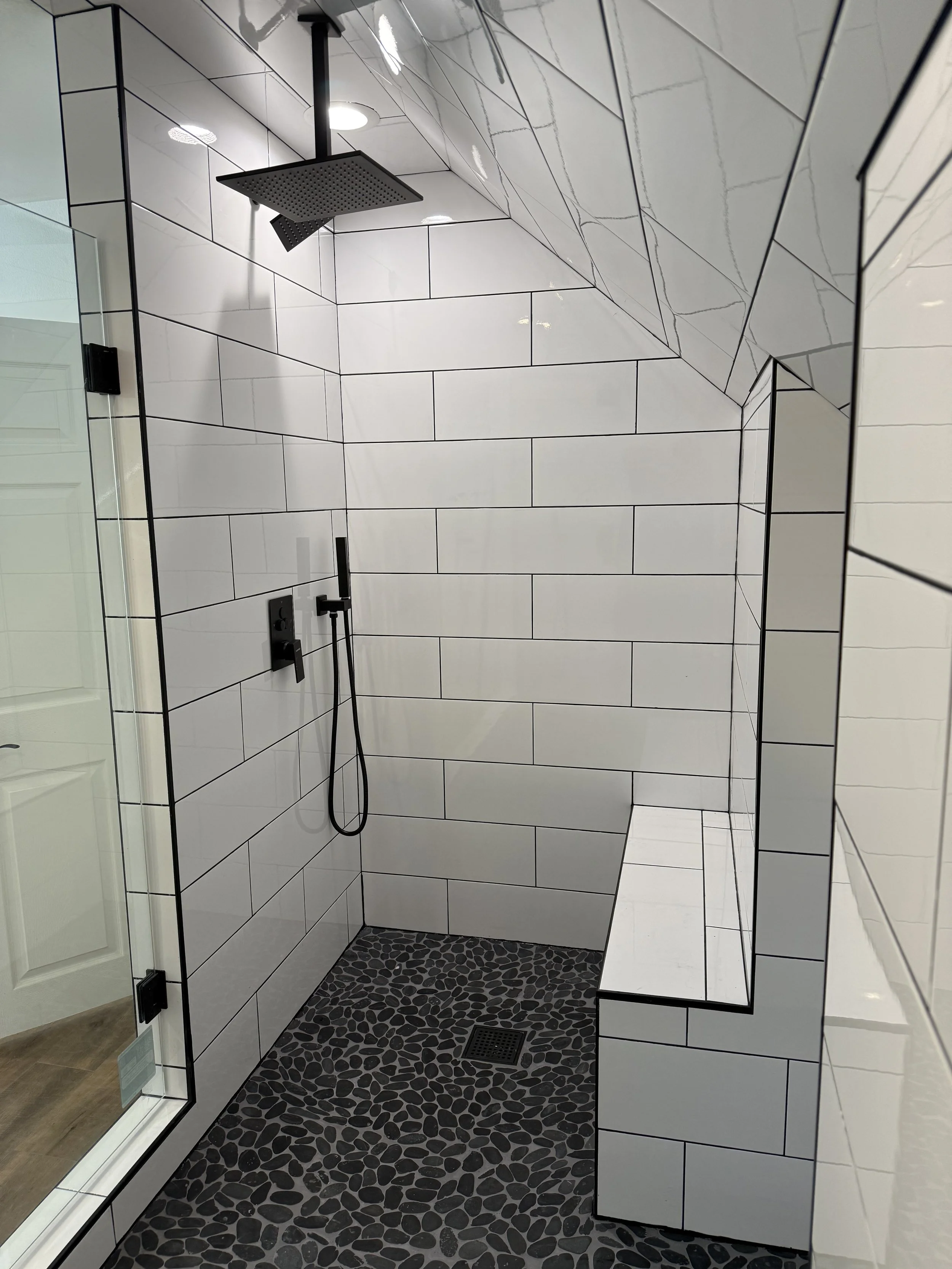 A modern walk-in shower with white subway tile walls and black grout, featuring a black rainfall showerhead, a black handheld shower, and a black floor drain with pebble-style black stones.