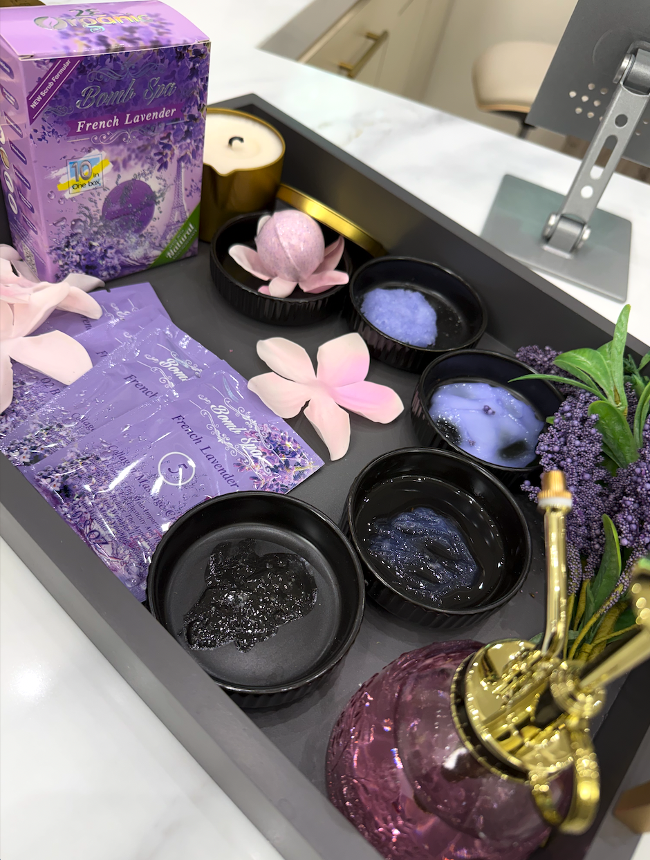A collection of purple and black candles, lavender sachets, artificial lavender flowers, pink flower petals, and a purple diffuser in a tray, with some tea lights and a purple and gold vase, all on a white countertop.