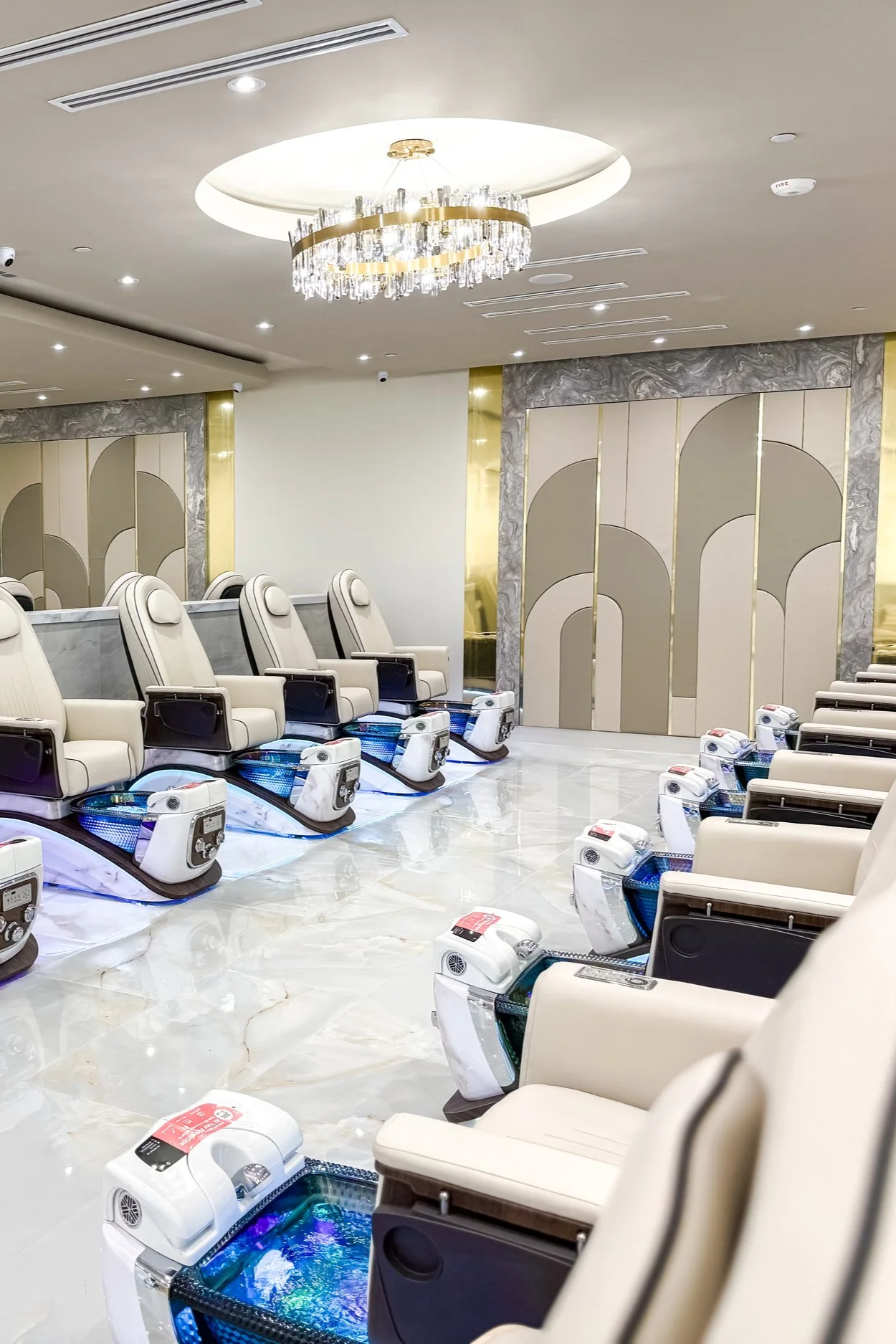 A shot of a luxury nail or spa salon with white massage chairs with built-in foot baths filled with water and colorful LED lights, arranged in rows in a spacious, well-lit room with elegant decor, marble floors, and a chandelier.