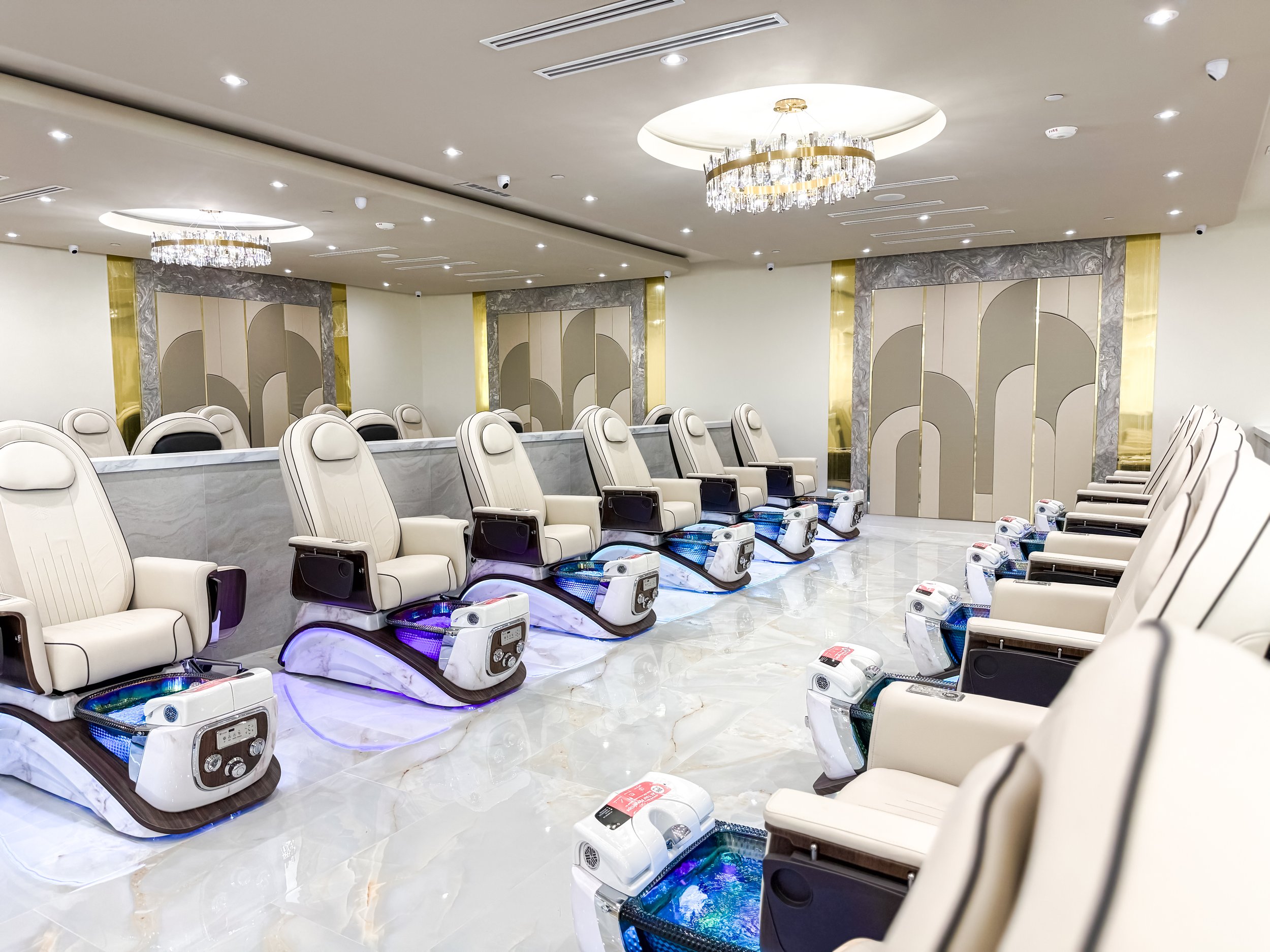 Luxury spa treatment room with rows of massage chairs equipped with foot baths, bright lighting, chandelier, marble accents, and elegant decor.