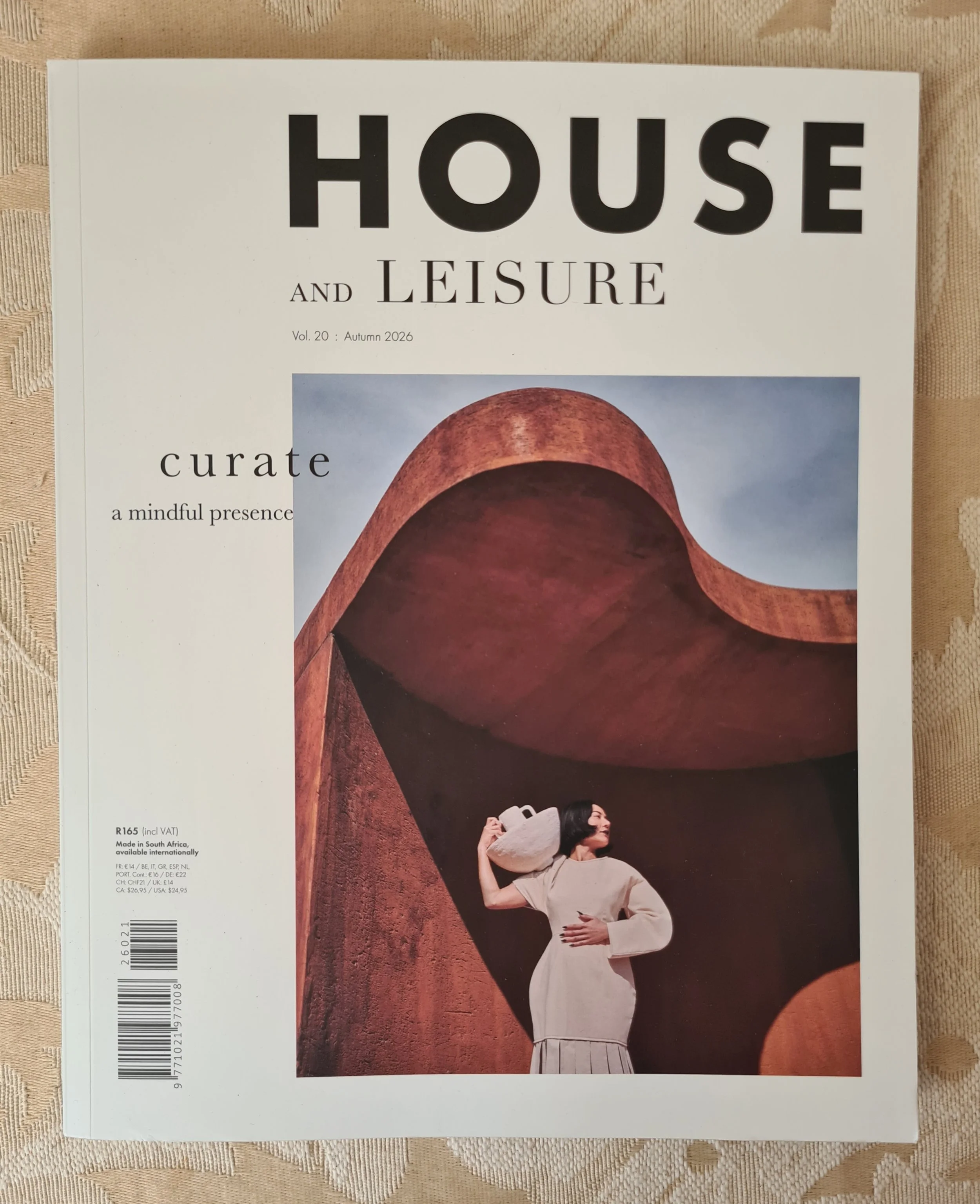 Magazine cover titled 'HOUSE AND LEISURE' Autumn 2026, featuring a woman in a white dress standing in front of a large curved architectural structure with a textured reddish-brown surface, holding a white bag.