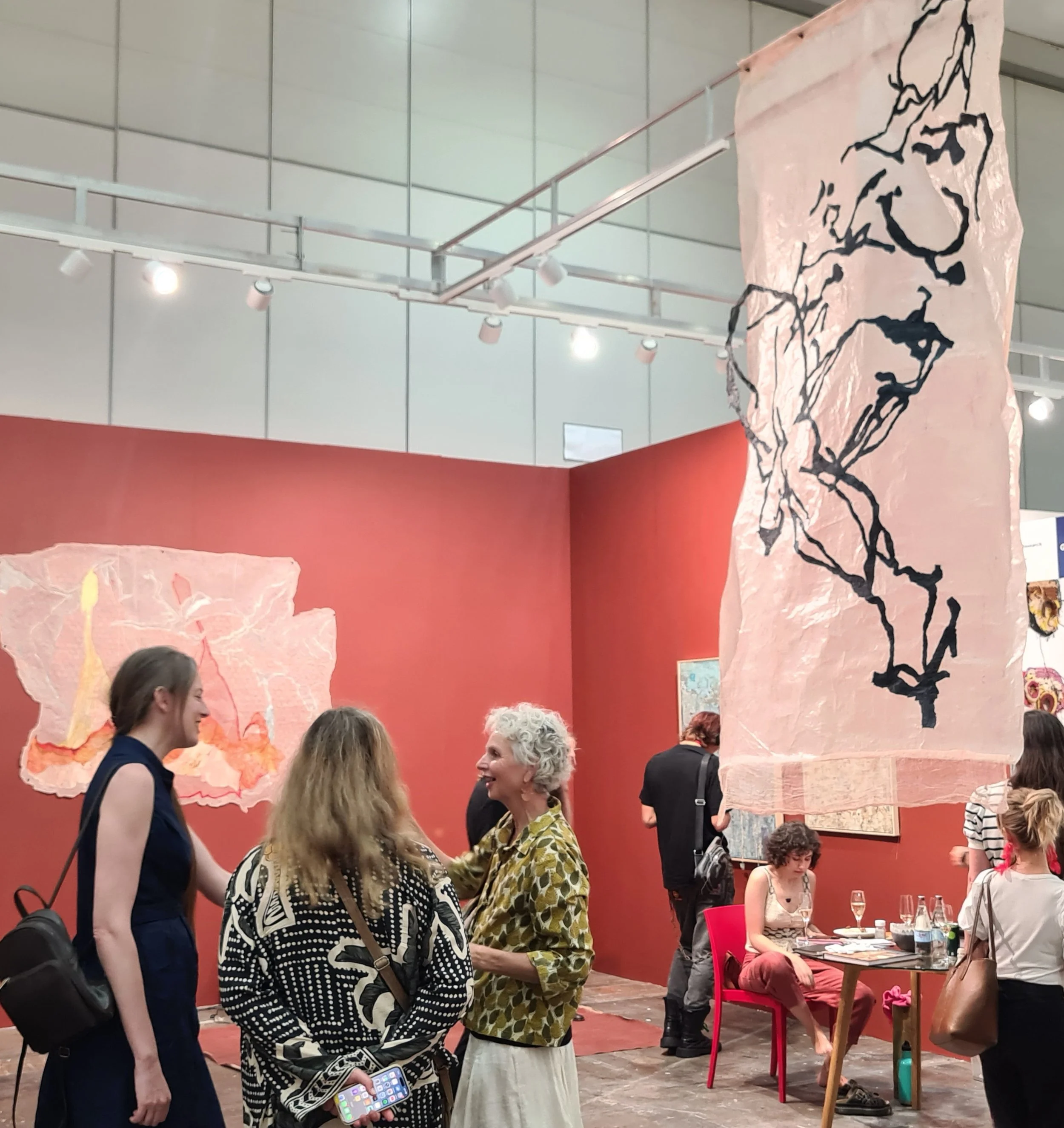 A group of women engaging in conversation at a social event in an indoor venue decorated with large pink and black artwork resembling a traditional Chinese ink painting. Some women are standing, while others are seated at a table with drinks.