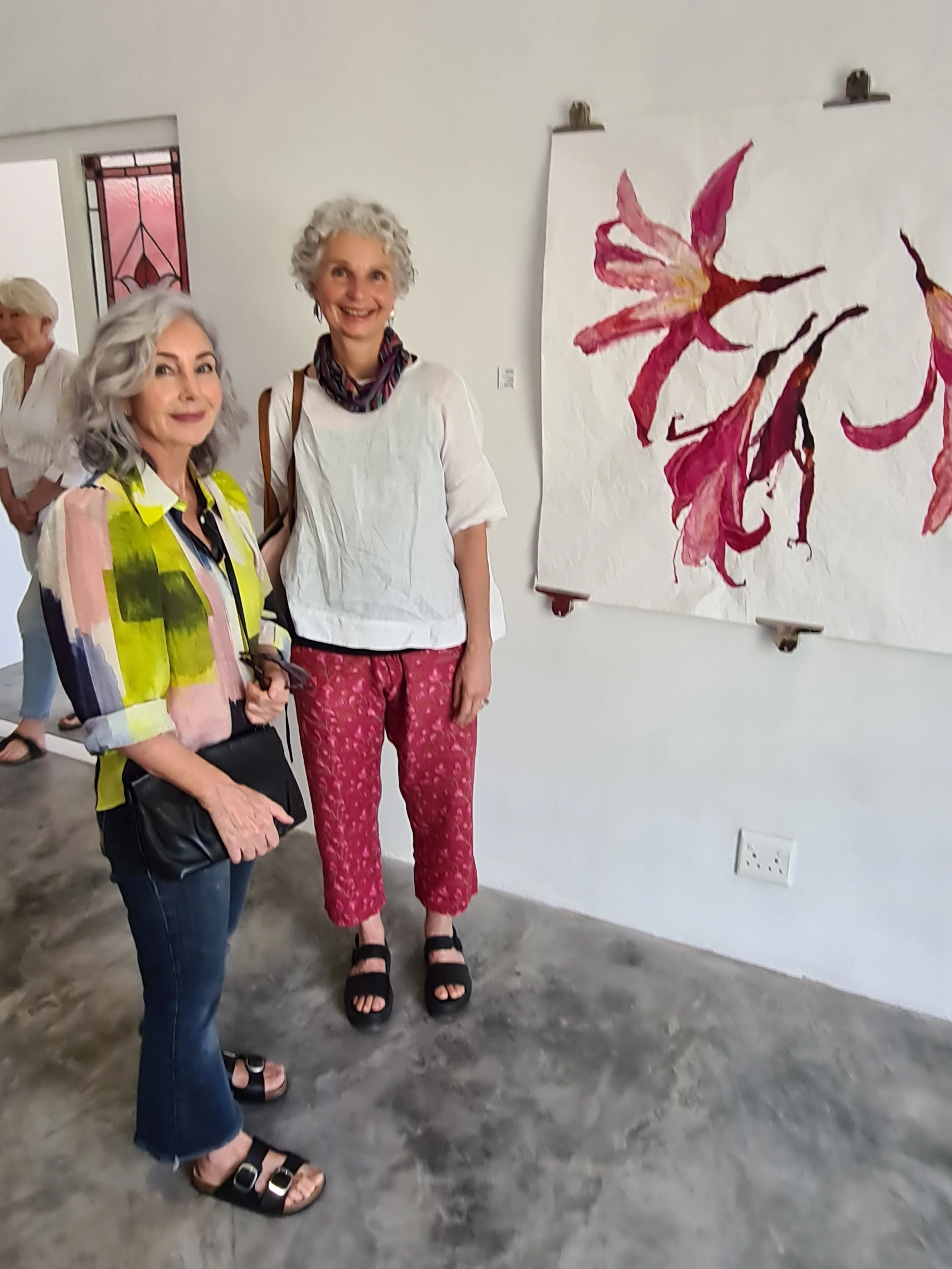 Two women standing next to each other in an art gallery, with a large watercolor painting of a pink and red flower hanging on the wall behind them.