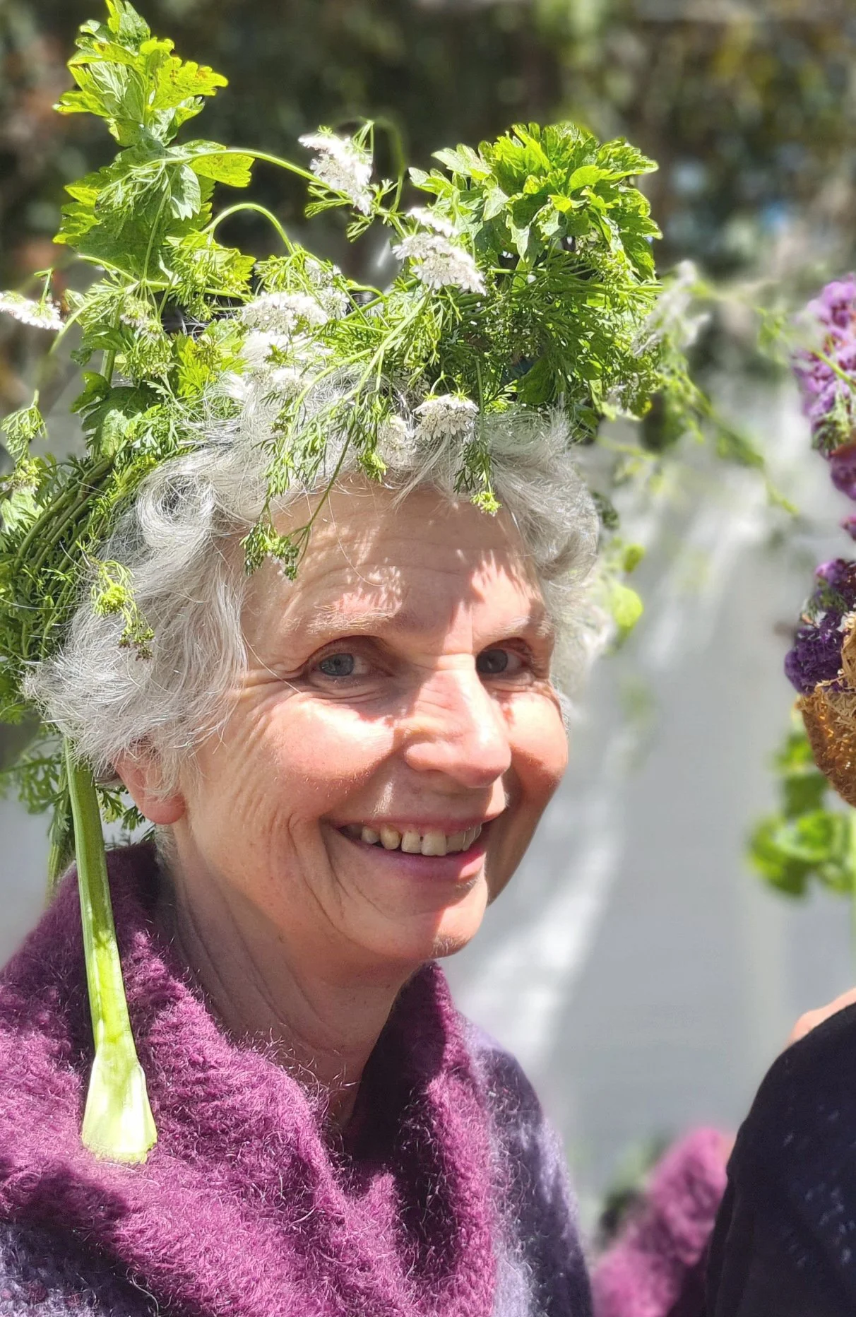 Smiling elderly woman wearing a purple fuzzy sweater, with a crown made of green leaves and white flowers on her head, outdoors on a sunny day.