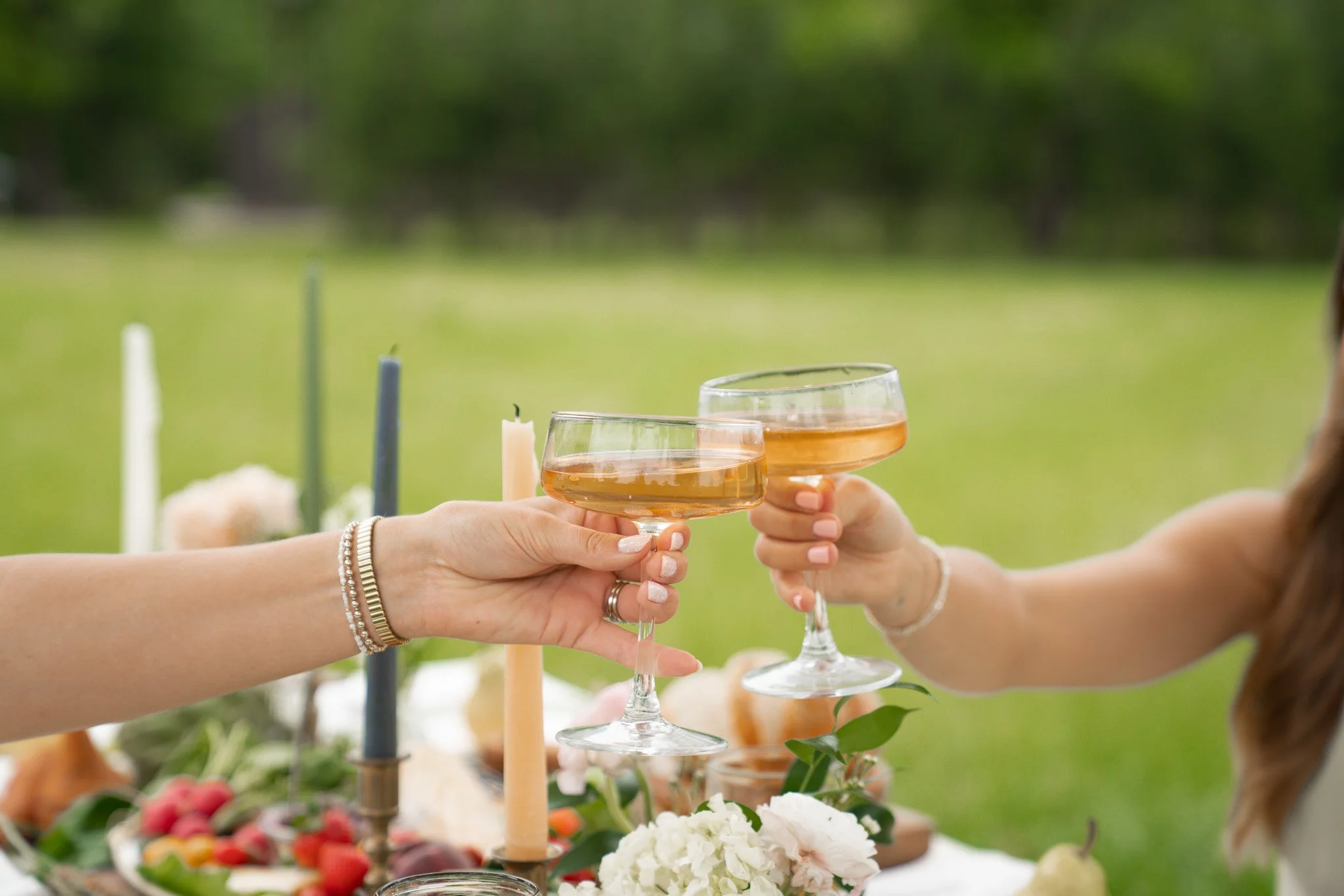 Two women raising glasses of rosé wine at an outdoor celebration with table of food and candles.