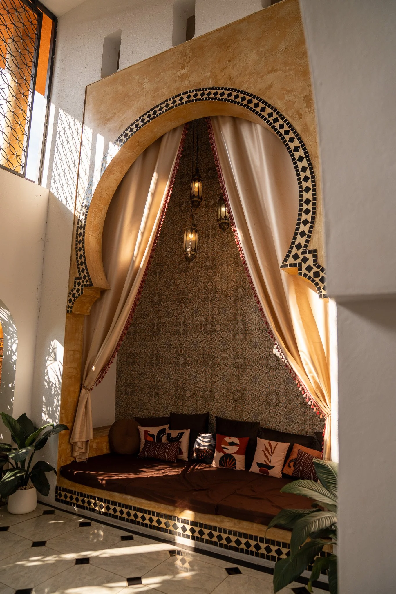 Interior of a room with Moroccan-style decor, featuring a curved ceiling with black and white mosaic tiles, beige drapes, and hanging lanterns. There is a cushioned seating area with decorative pillows and greenery on the side.