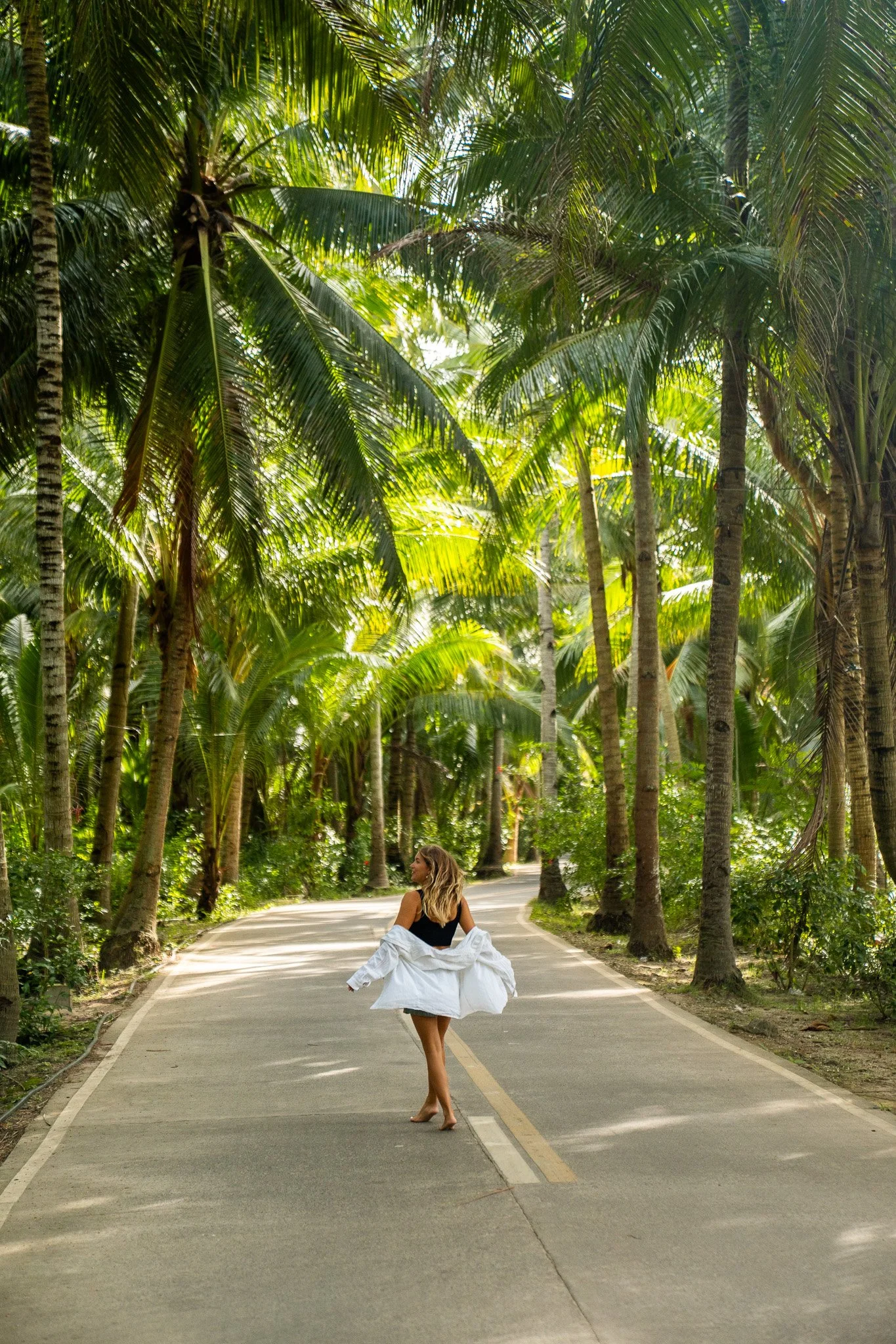A woman with blonde hair, wearing a black top and a white jacket, is standing on a winding road surrounded by tall palm trees, with lush green foliage. She appears to be walking or dancing on the road.