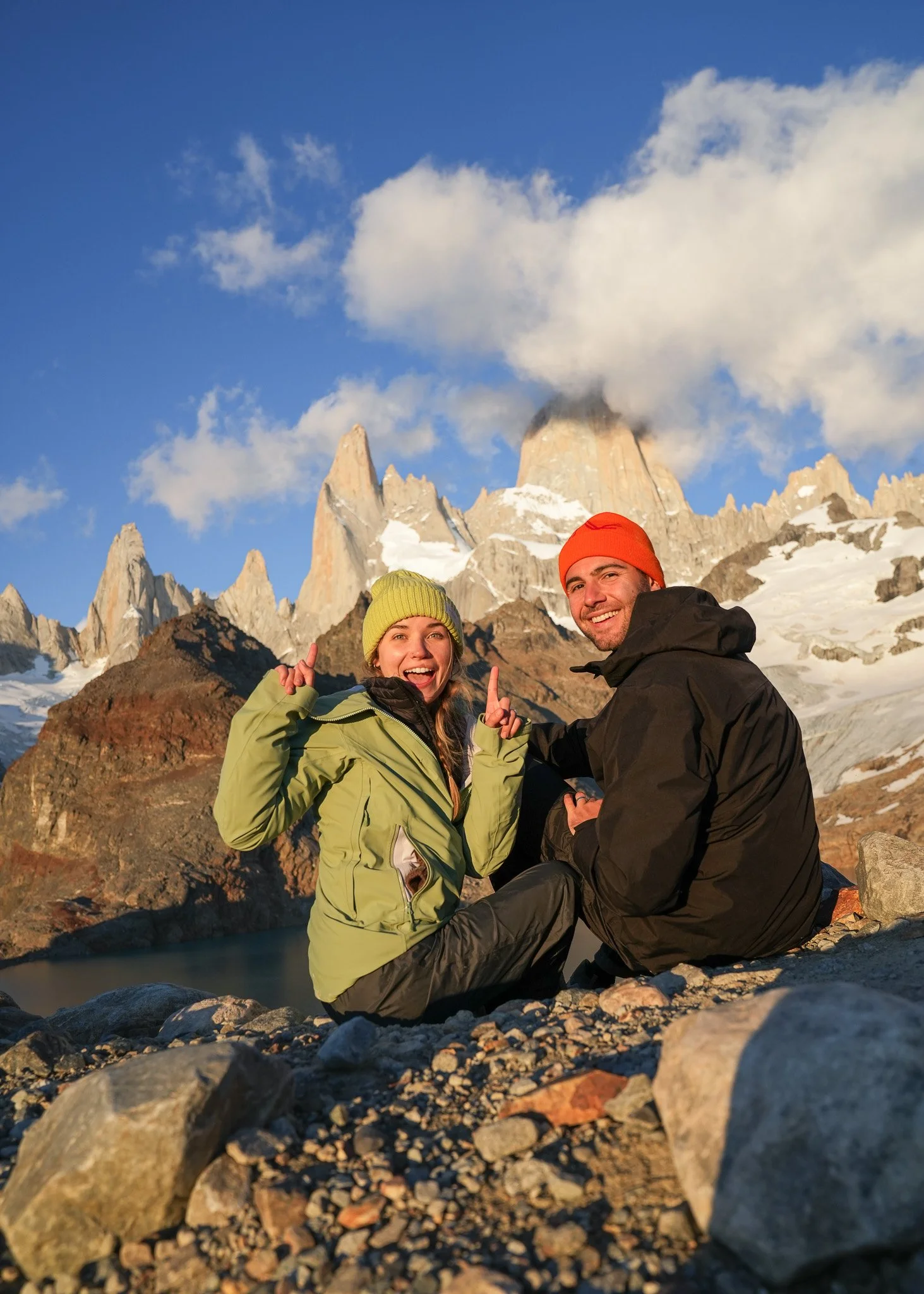Two hikers, a woman and a man, sitting on rocky terrain in front of a mountain range with snow and clouds in the background. The woman is wearing a green jacket and yellow beanie, smiling and pointing upward, while the man is wearing a black jacket and red beanie, smiling at the camera.