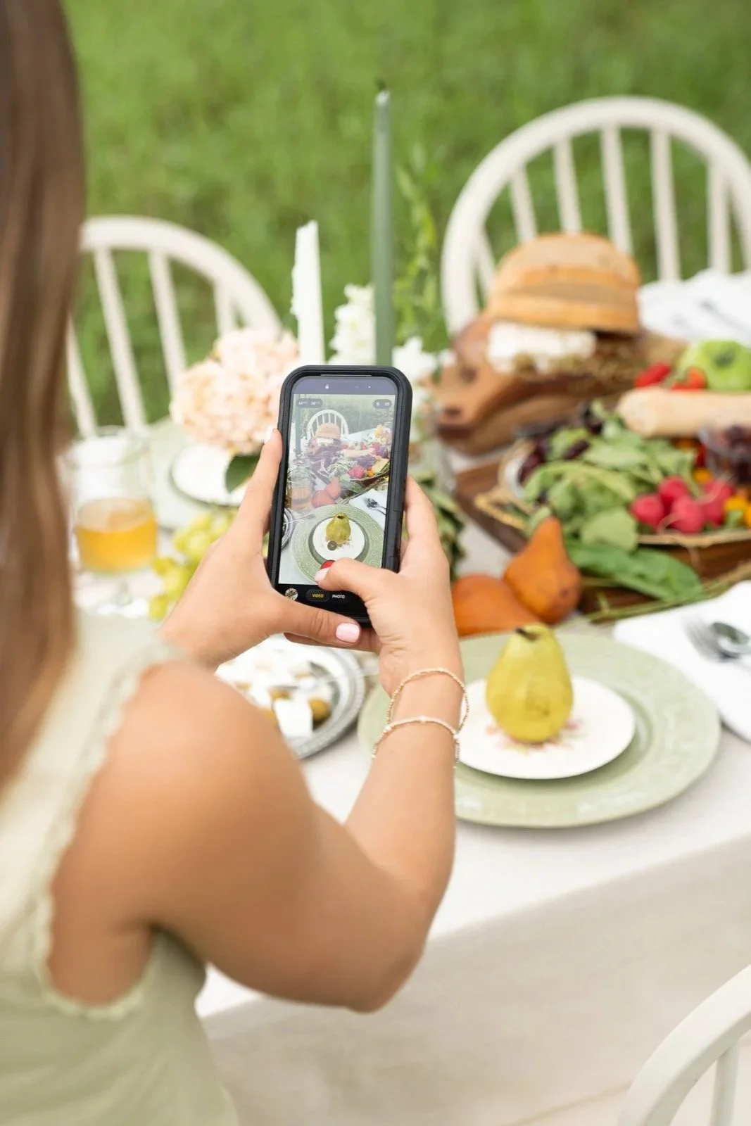 Person taking a photo of a table setting with a smartphone. The table has various foods, including a pear, bread, vegetables, and a drink, with white chairs and a green outdoor background.
