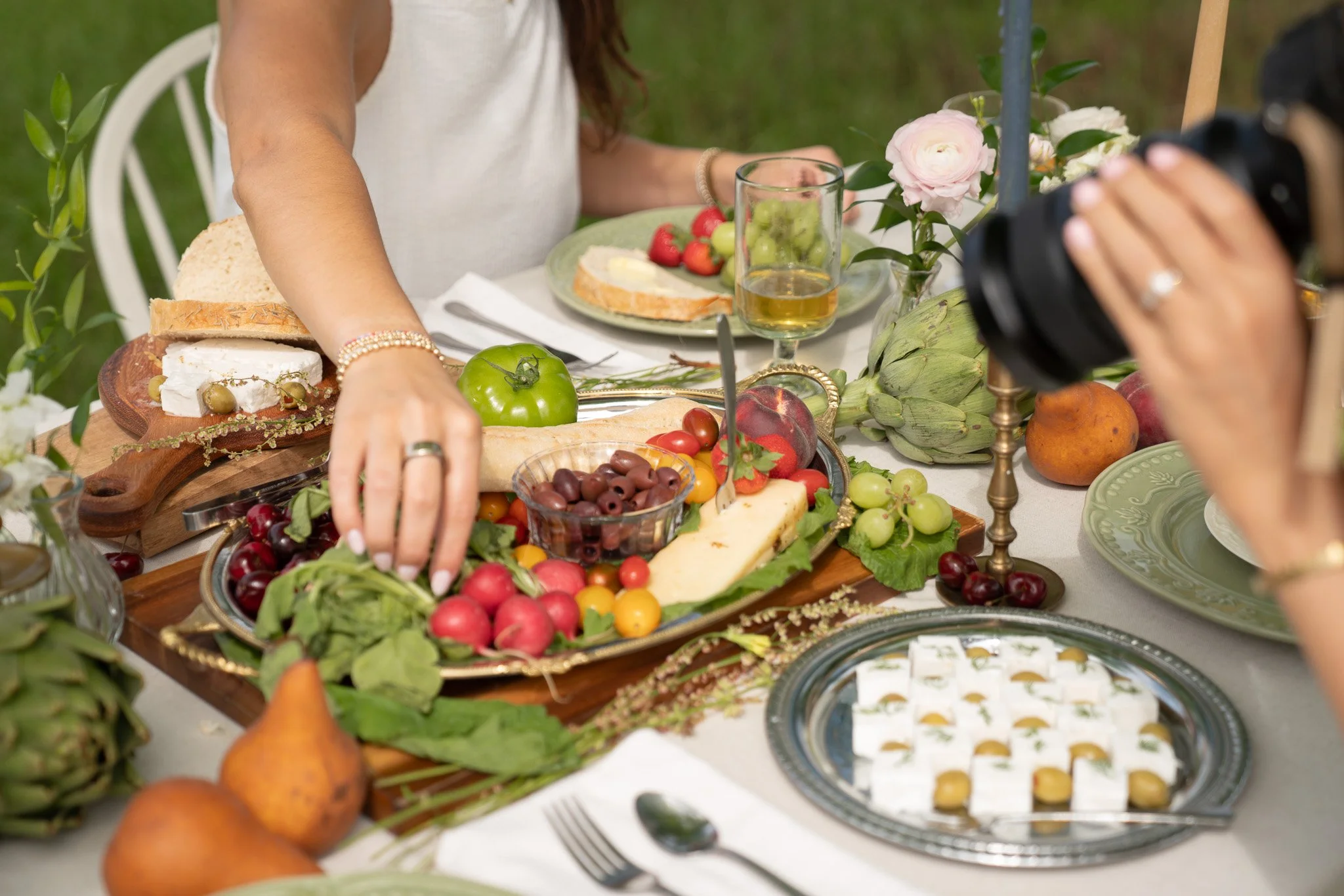 A woman arranging a table filled with various foods including cheese, bread, strawberries, cherries, grapes, and artichokes, with flowers and glasses of drink on a decorated outdoor table.