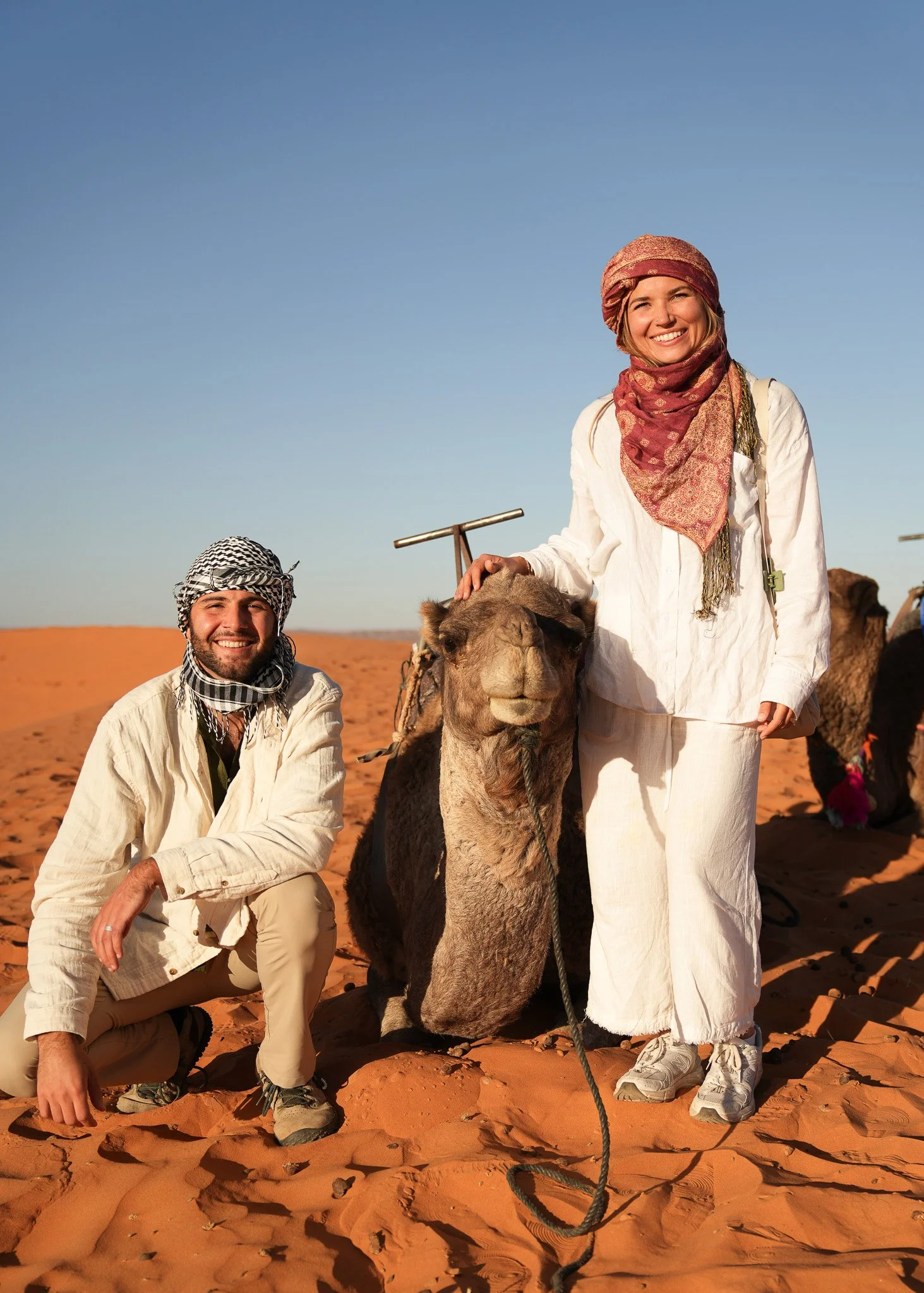 Two smiling tourists in desert attire with a camel in a sandy desert landscape under a clear blue sky.