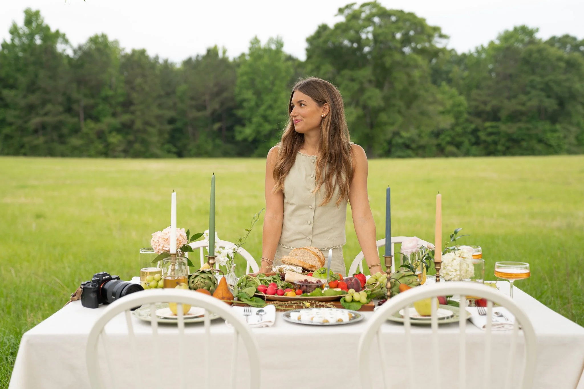 A woman with brown hair and pearl earrings standing behind a decorated table outdoors in a field with green trees in the background. The table is set with food, flowers, candles, and a camera.
