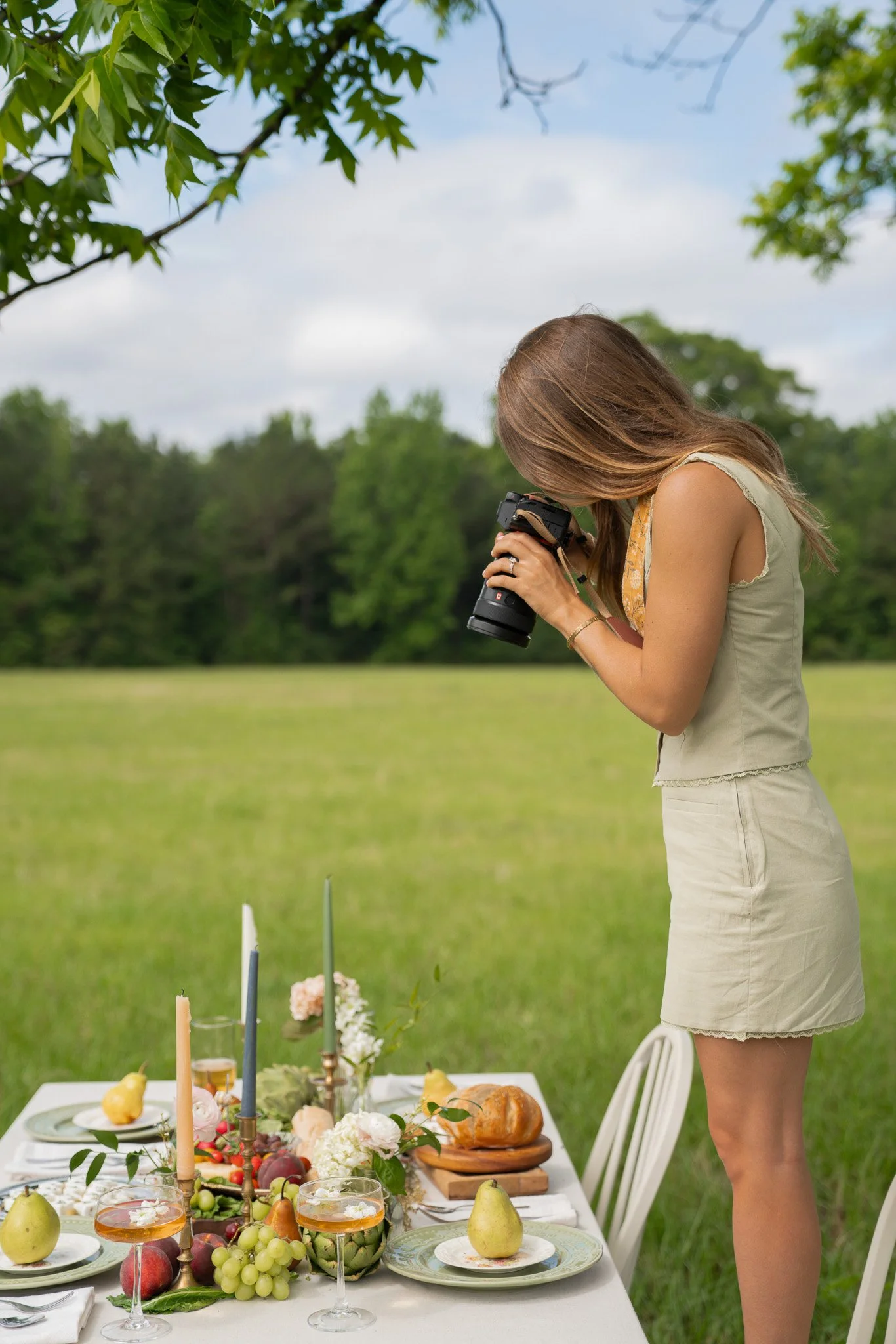 A woman with long brown hair wearing a beige dress standing in a grassy outdoor setting, preparing or setting a table with food for a gathering or celebration, with green trees and a cloudy sky in the background.