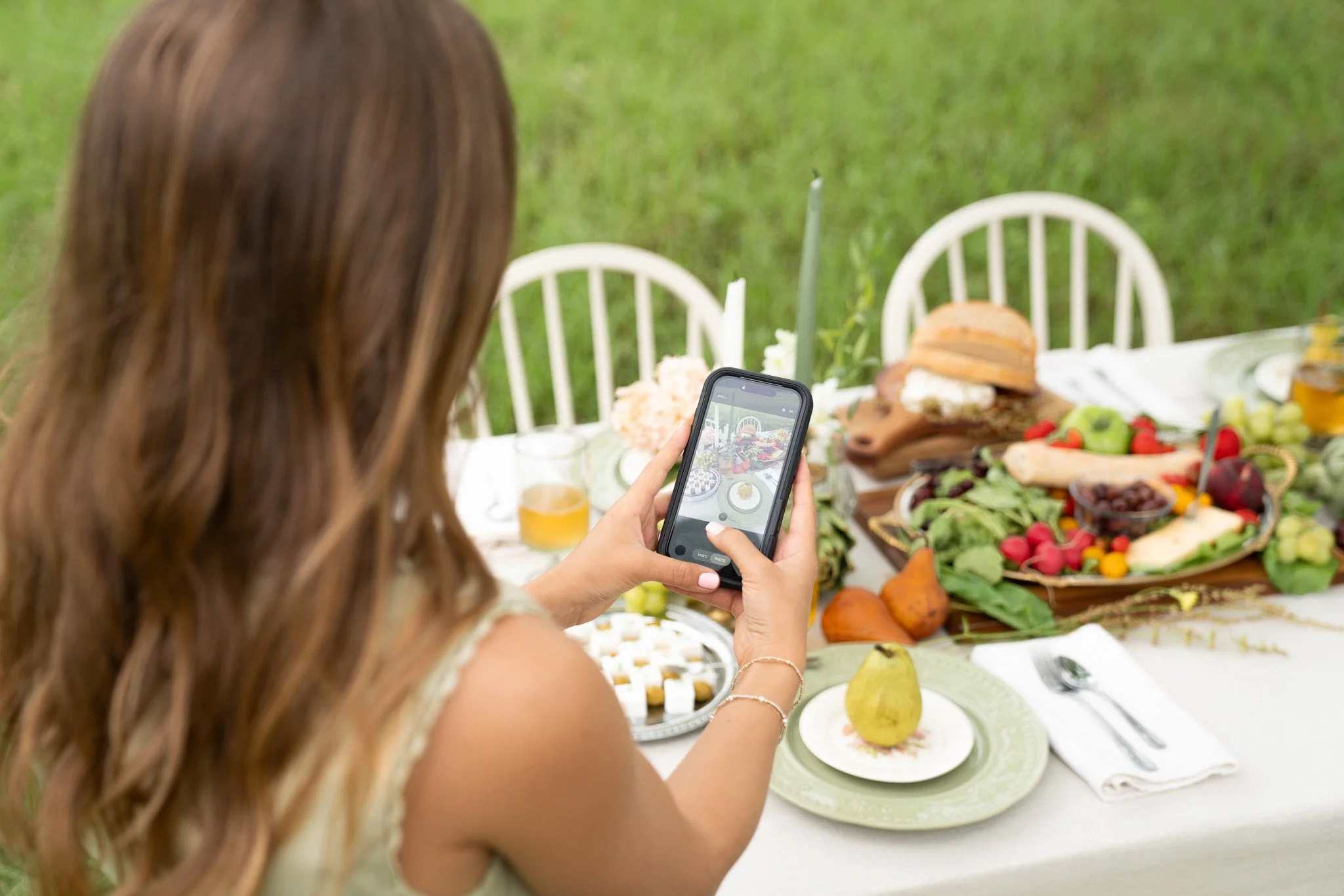A woman with long brown hair taking a photo with her phone of an outdoor table set with various foods including bread, fruits, and vegetables, on a grassy background.
