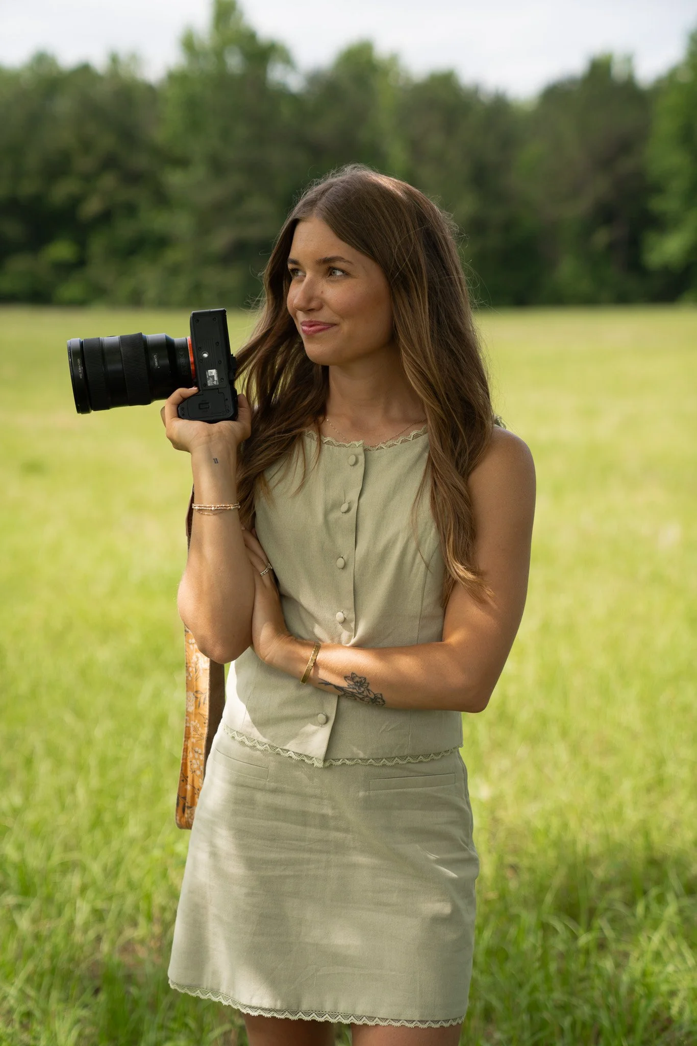 A woman standing in a field with grass, holding a camera with a large lens on her shoulder, looking to her right with a slight smile.