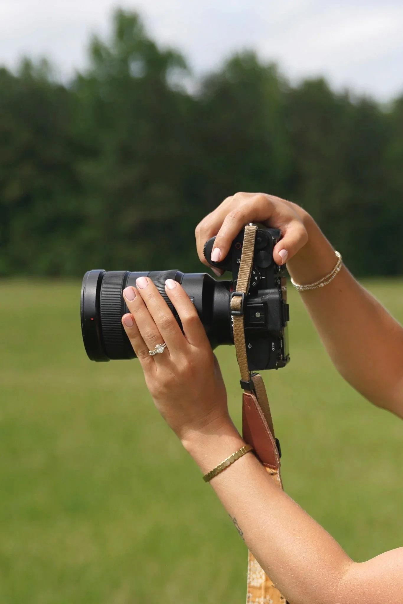 Person holding a professional digital camera outdoors in a grassy field with trees in the background.