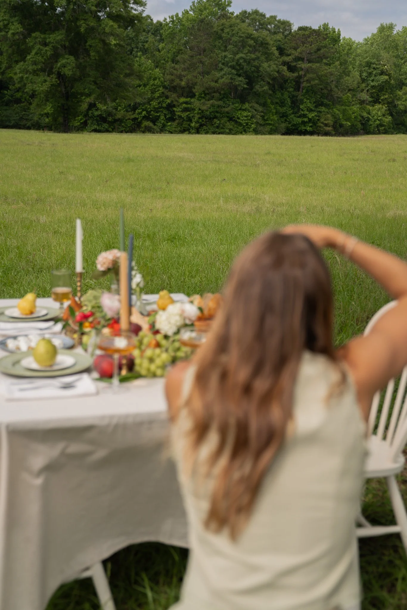 Woman with long hair sitting at a table outdoors in a grassy field, with a backdrop of trees and cloudy sky.