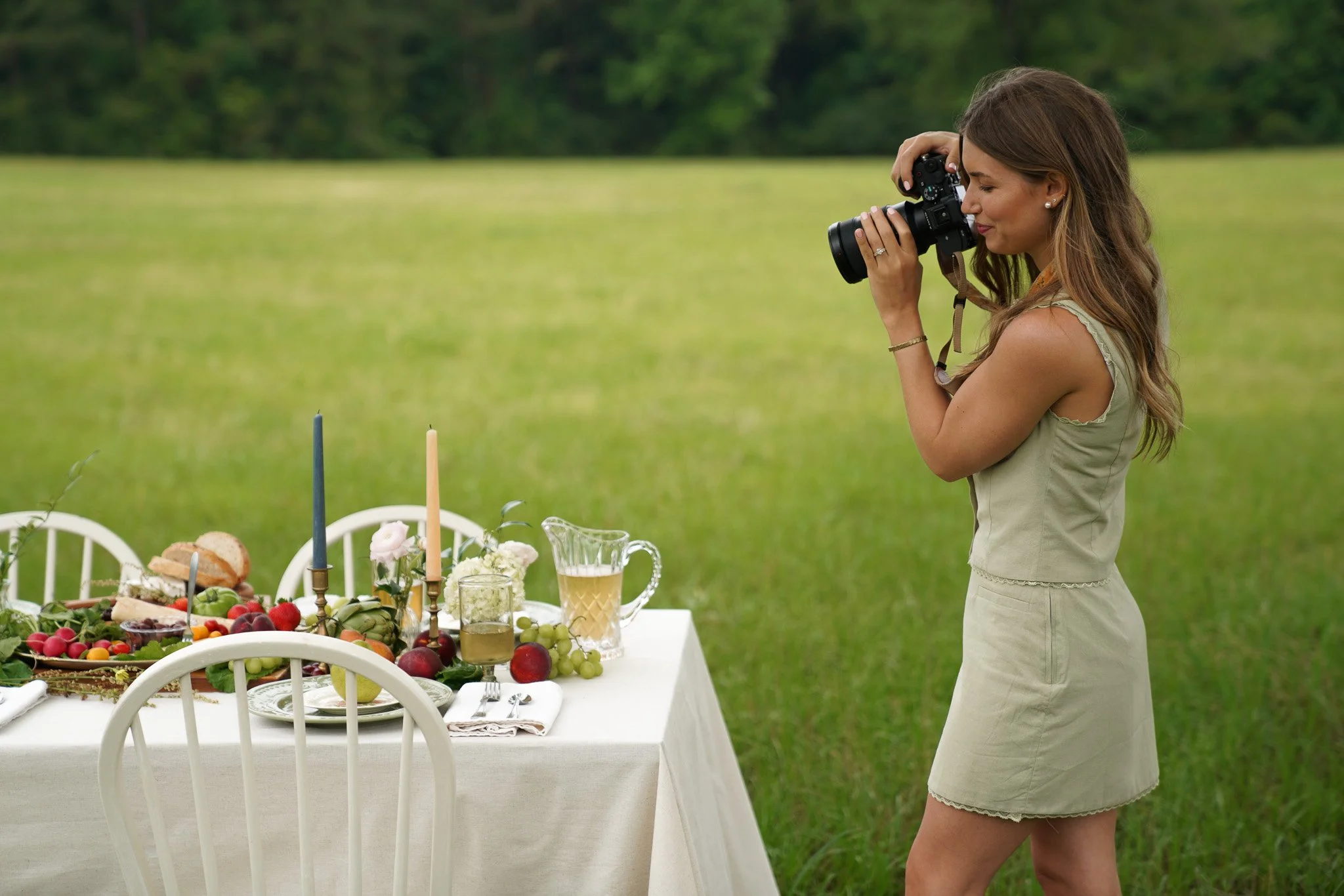 A woman with long brown hair, wearing a sleeveless dress, takes a photograph at an outdoor gathering in a green field with a decorated table of food and drinks.