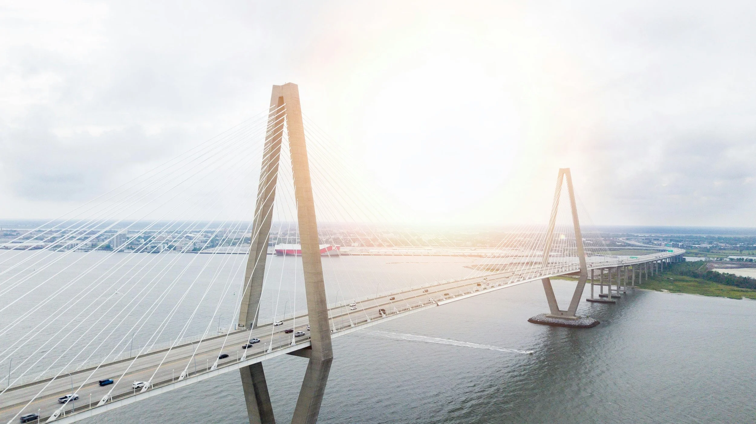 A large cable-stayed bridge with two main pylons crosses a wide river, with cars traveling on the roadway and a boat in the water underneath. The sky is partly cloudy with sunlight shining through.