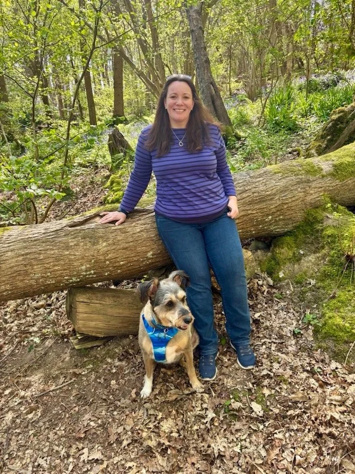 A woman in a blue striped long sleeve shirt and jeans sitting on a fallen tree trunk in a lush green forest, with a brown and white dog wearing a blue vest sitting beside her.