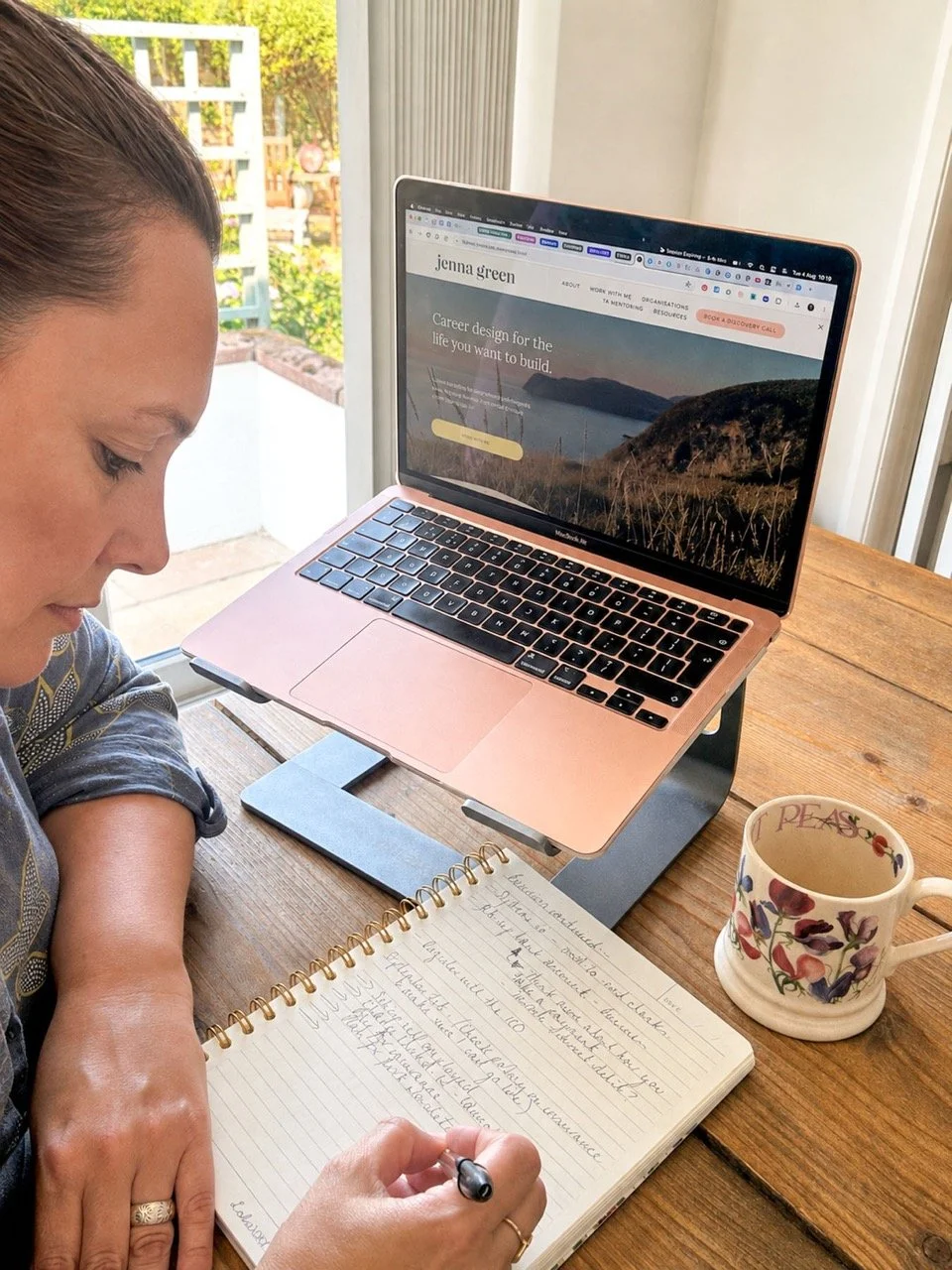 Woman writing in a notebook at a wooden table with a laptop, a colorful coffee mug, and a window with a garden view in the background.