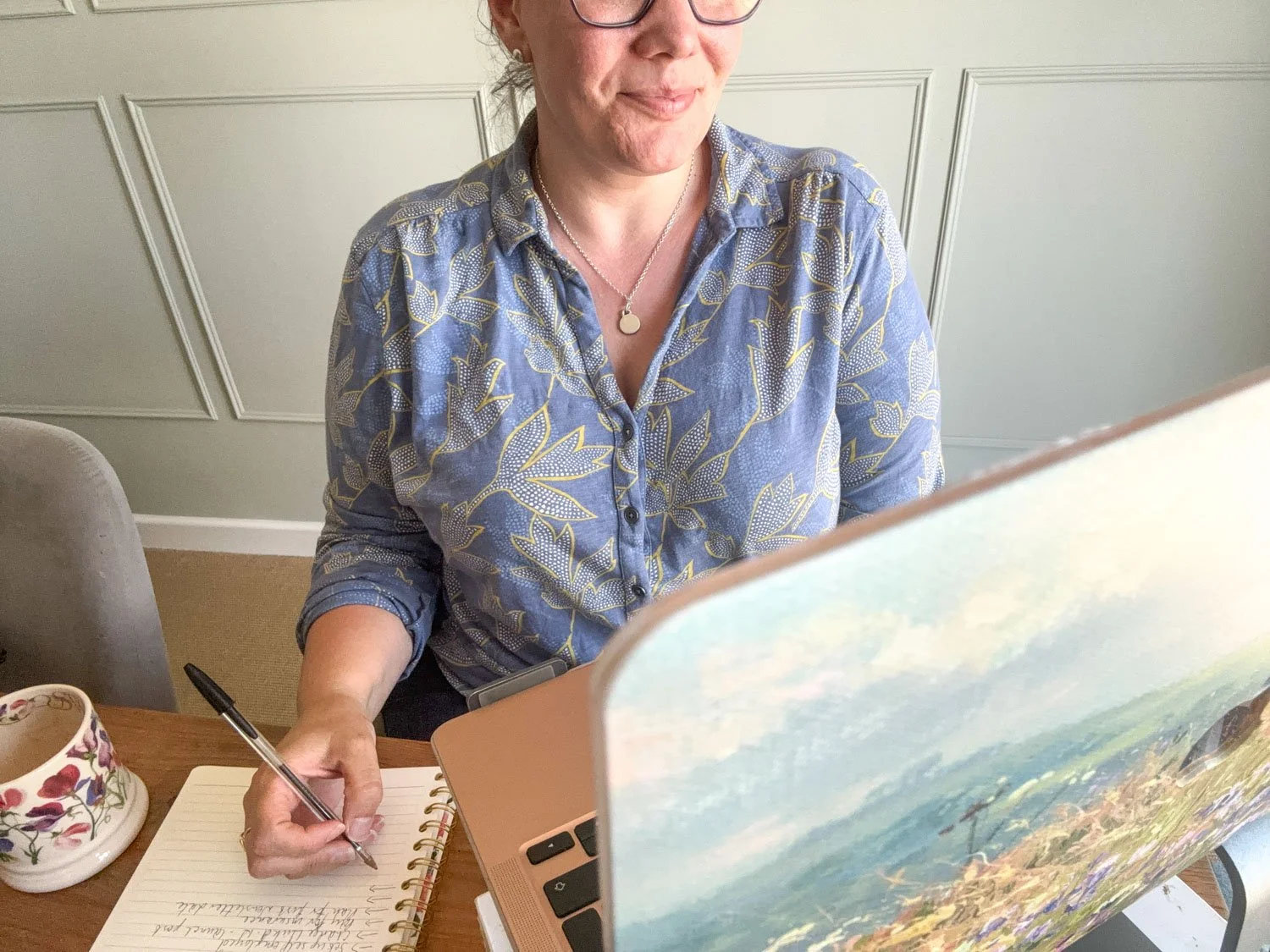 A woman wearing glasses and a patterned blue shirt is sitting at a table, writing in a spiral notebook with a black pen. She is partially obscured by a laptop displaying a scenic landscape image. There is a floral designed cup or mug on the table.