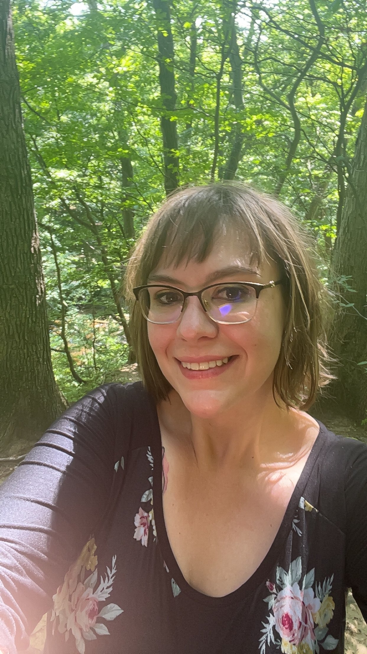 An image of Brittany outdoors in a wooded area with green leaves and trees in the background. She is wearing glasses, a black top with a floral pattern, and has shoulder-length brown hair.