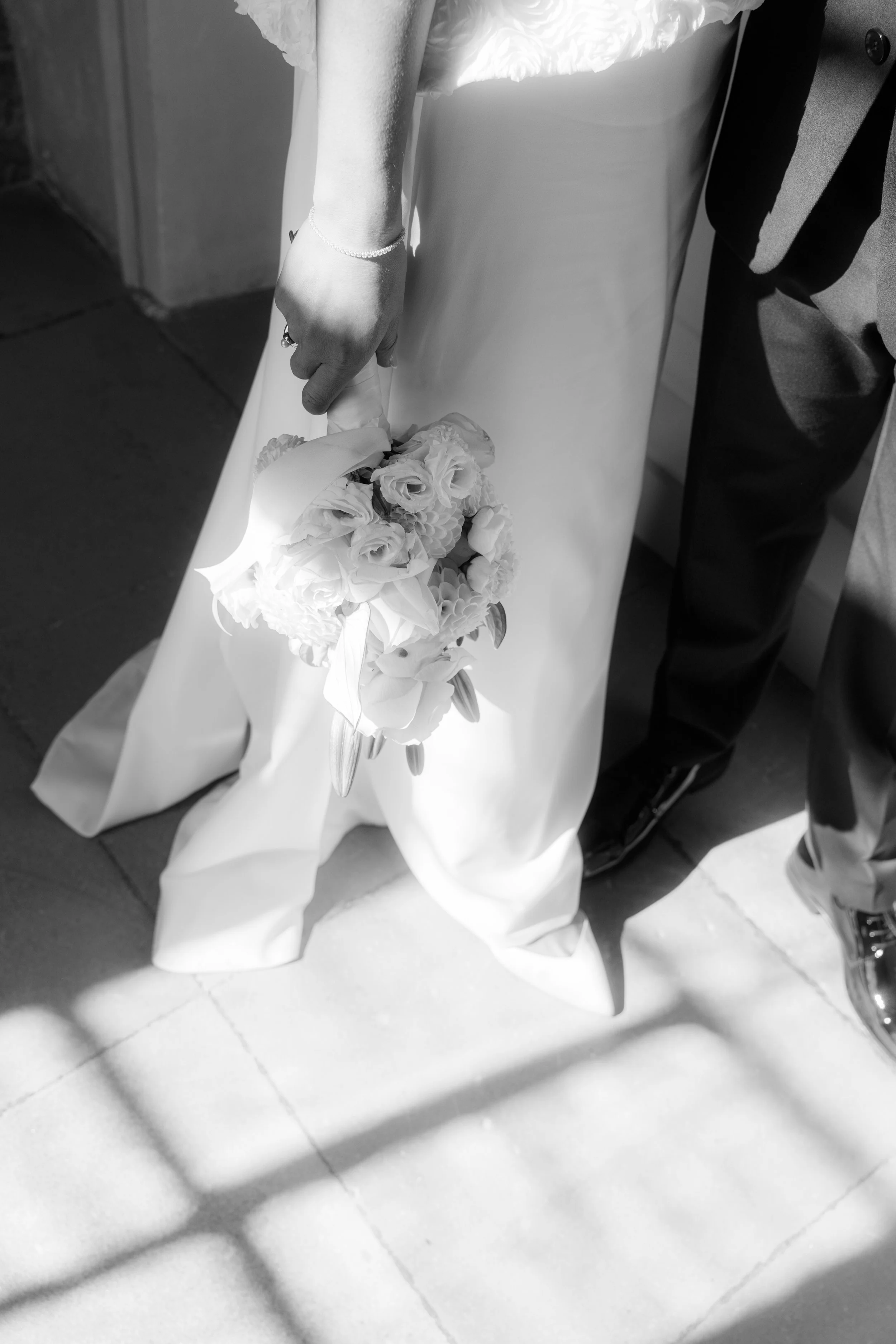 A person wearing a formal suit and dress shoes holding a bouquet of flowers, with their hand resting on a table or surface.