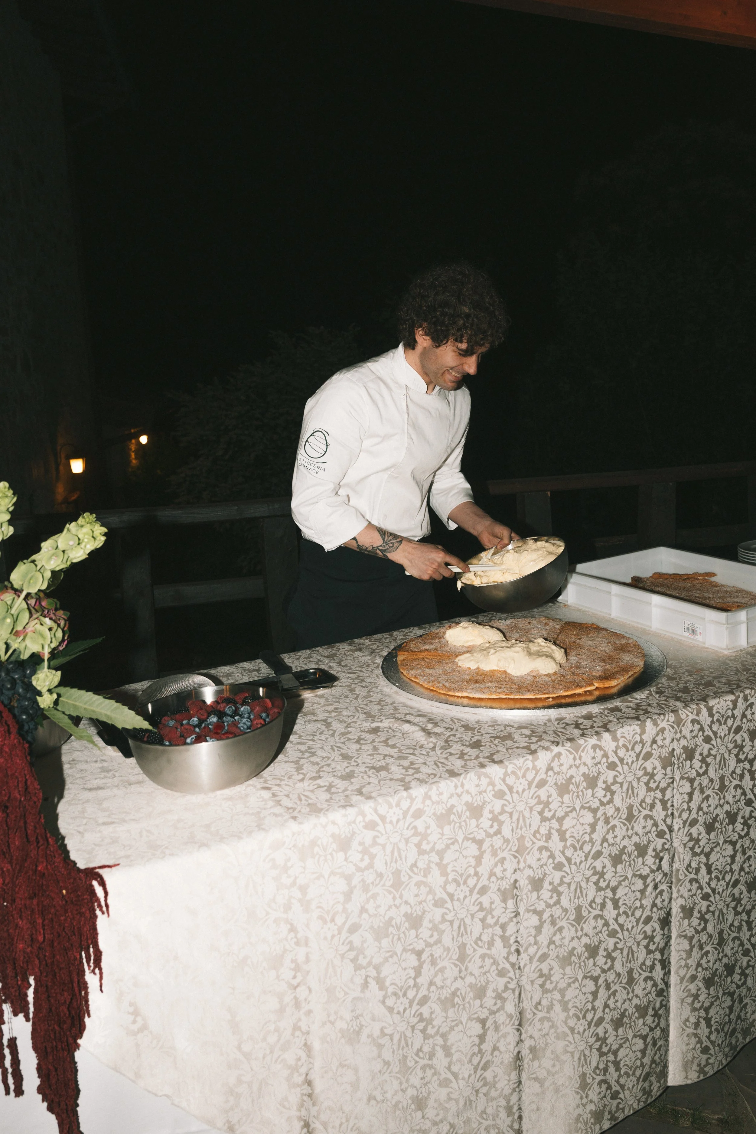 A chef in a white uniform preparing food at a table with a patterned tablecloth during an outdoor event at night. The table has bowls of berries and a large pizza crust with toppings.