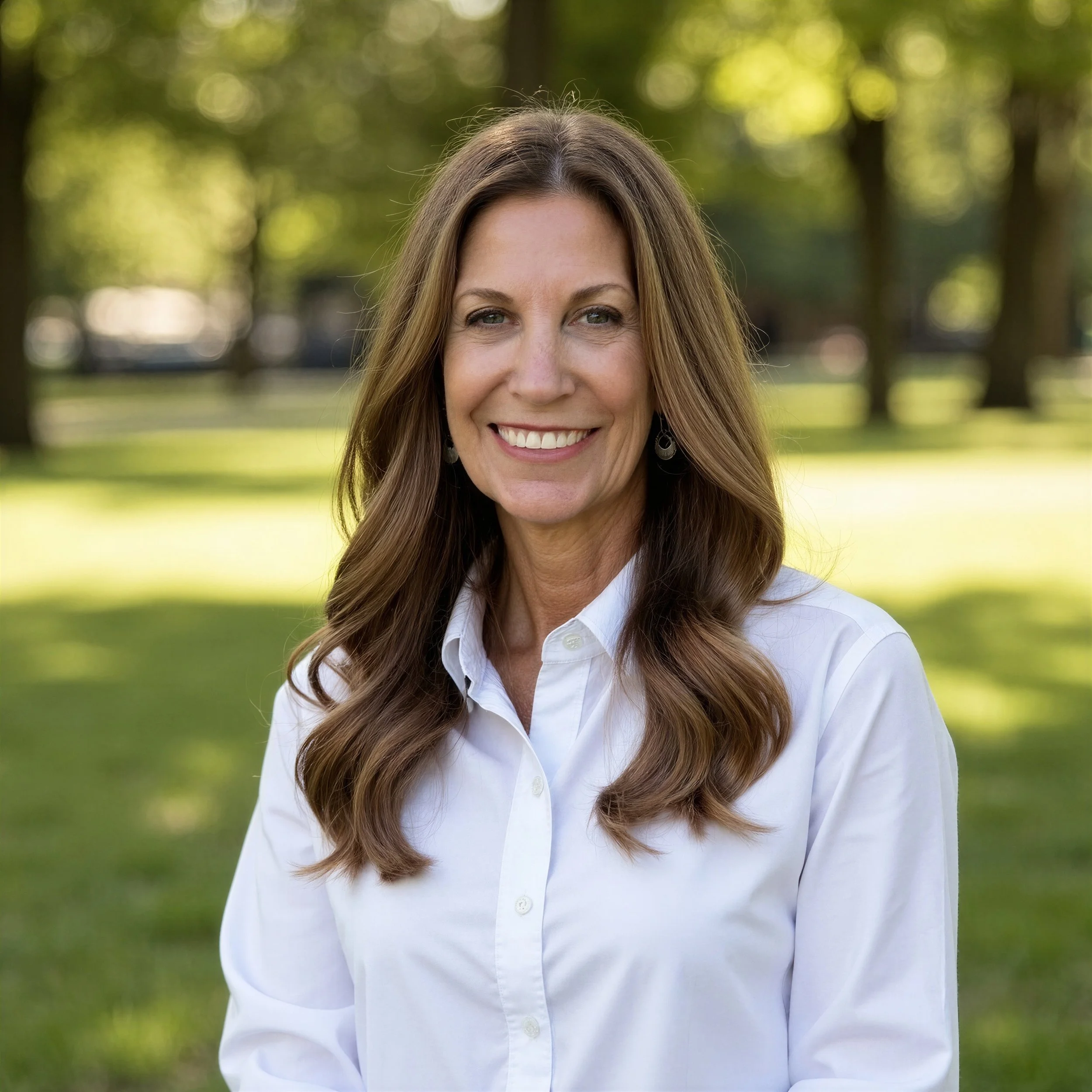 A smiling woman with long brown hair in a white shirt standing outdoors in a park with green trees and grass in the background.
