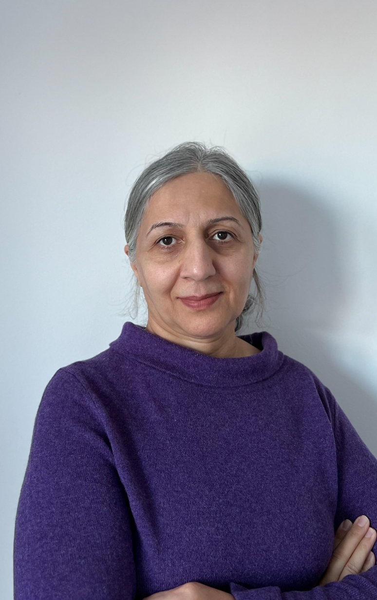A woman with gray hair wearing a purple sweater crossing her arms and standing against a plain white wall.